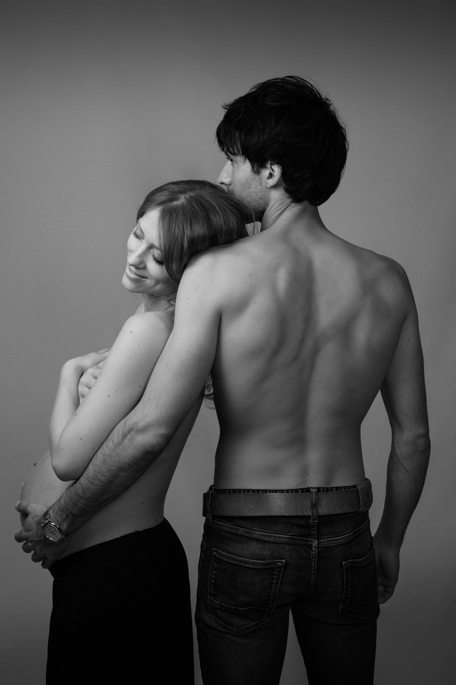 Black and white maternity portrait of a shirtless man embracing a pregnant woman from behind in a studio.