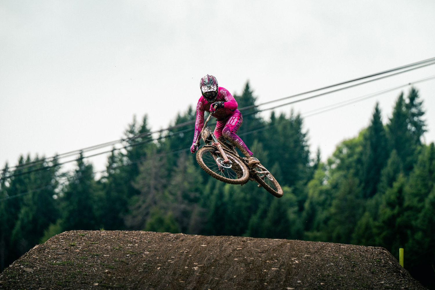 Mountain biker in pink gear catching big air off a dirt jump with forest in background.