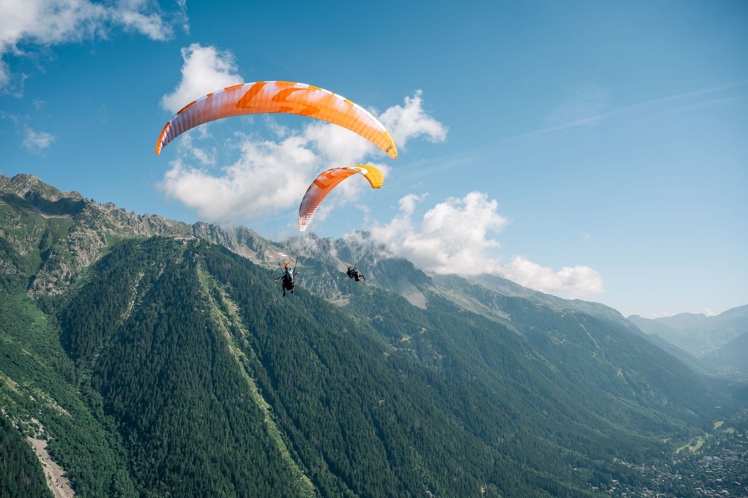 Two orange paragliders flying high above a scenic alpine valley with mountains and clouds.