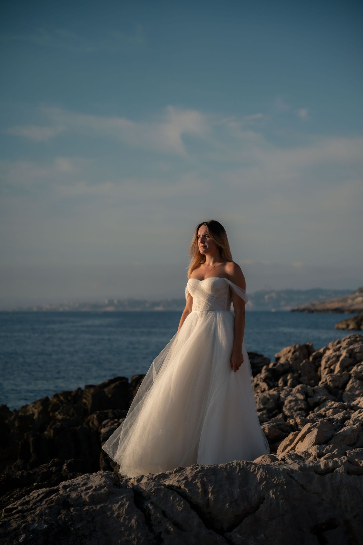 Bride in white off-shoulder tulle gown standing on rocky coastal shore with blue sea and sky backdrop.