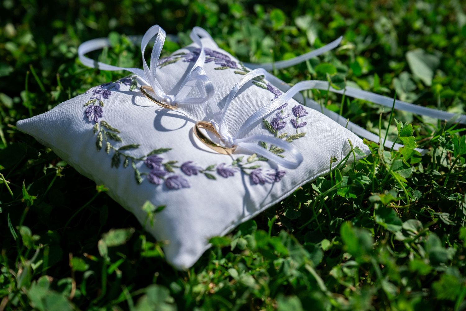 White ring bearer pillow with lavender embroidery and wedding rings resting on green grass.