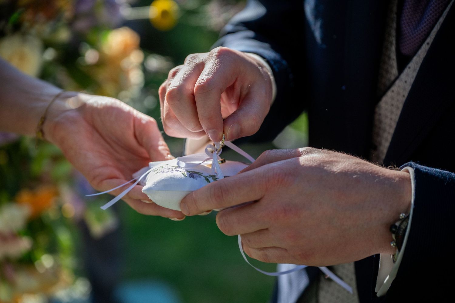 Close-up of hands exchanging wedding rings on a white pillow during an outdoor ceremony.