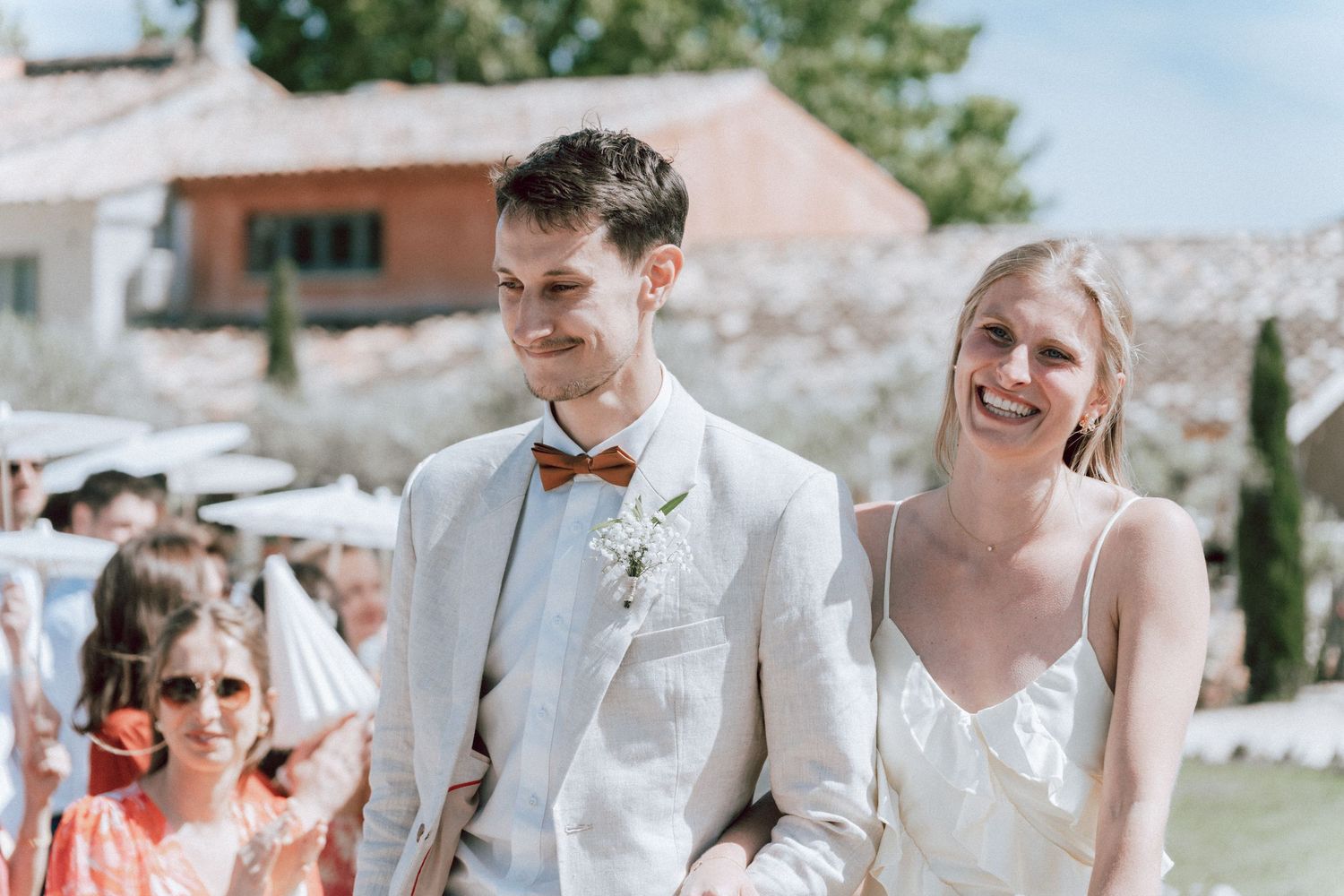 Smiling bride and groom in linen suit with orange bow tie walking at outdoor wedding.