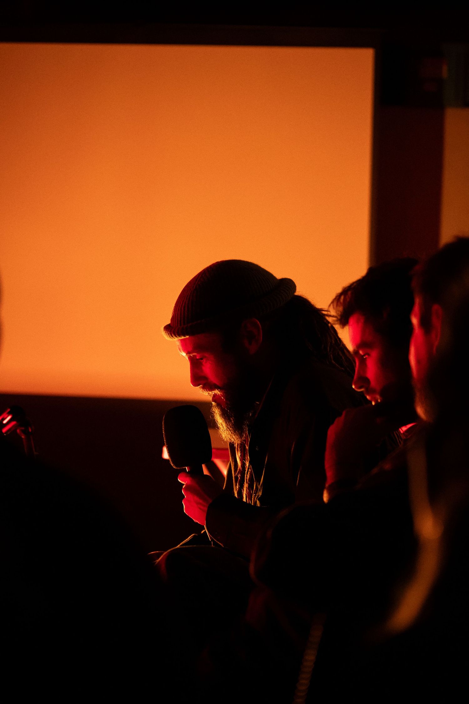 Bearded man holding a microphone in warm orange light at an indoor event.