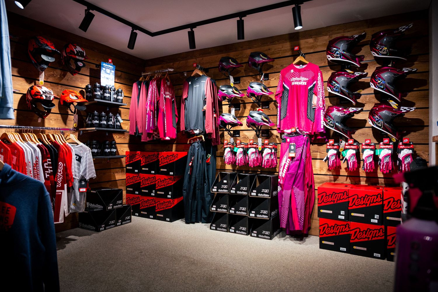 Retail sports store displaying pink and red cycling gear, helmets, and apparel on wooden wall-mounted racks.