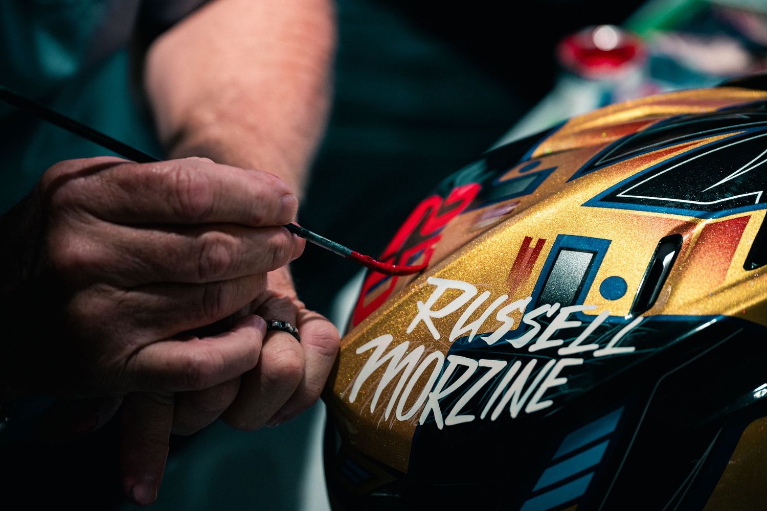 A hand carefully paints 'Russell Morzine' lettering onto a colorful racing helmet using a fine brush.