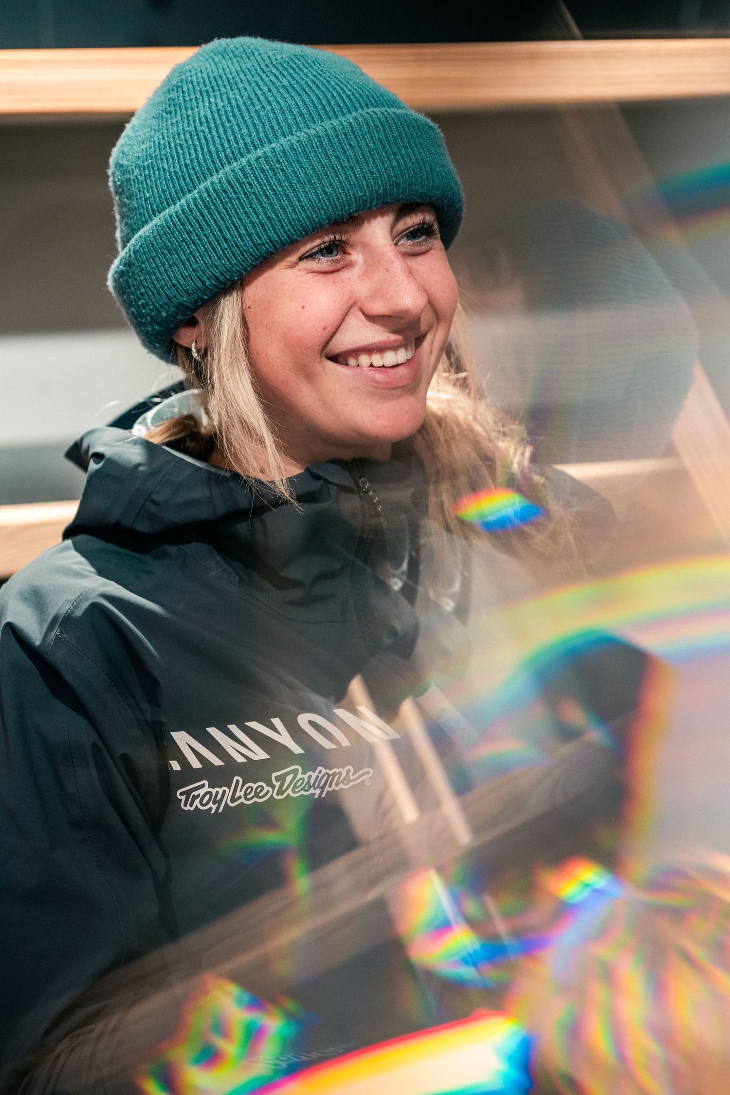 Smiling woman wearing a teal beanie and Canyon Troy Lee Designs jacket, with colorful light refractions in the foreground.