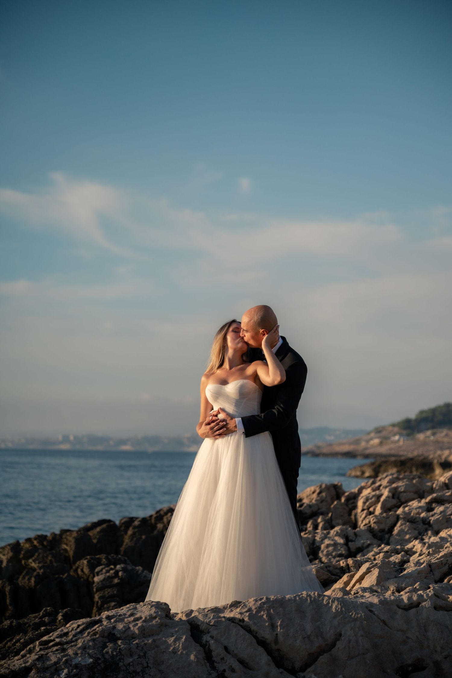 Bride and groom kissing on rocky seaside at golden hour, bride in white tulle gown, groom in dark suit.