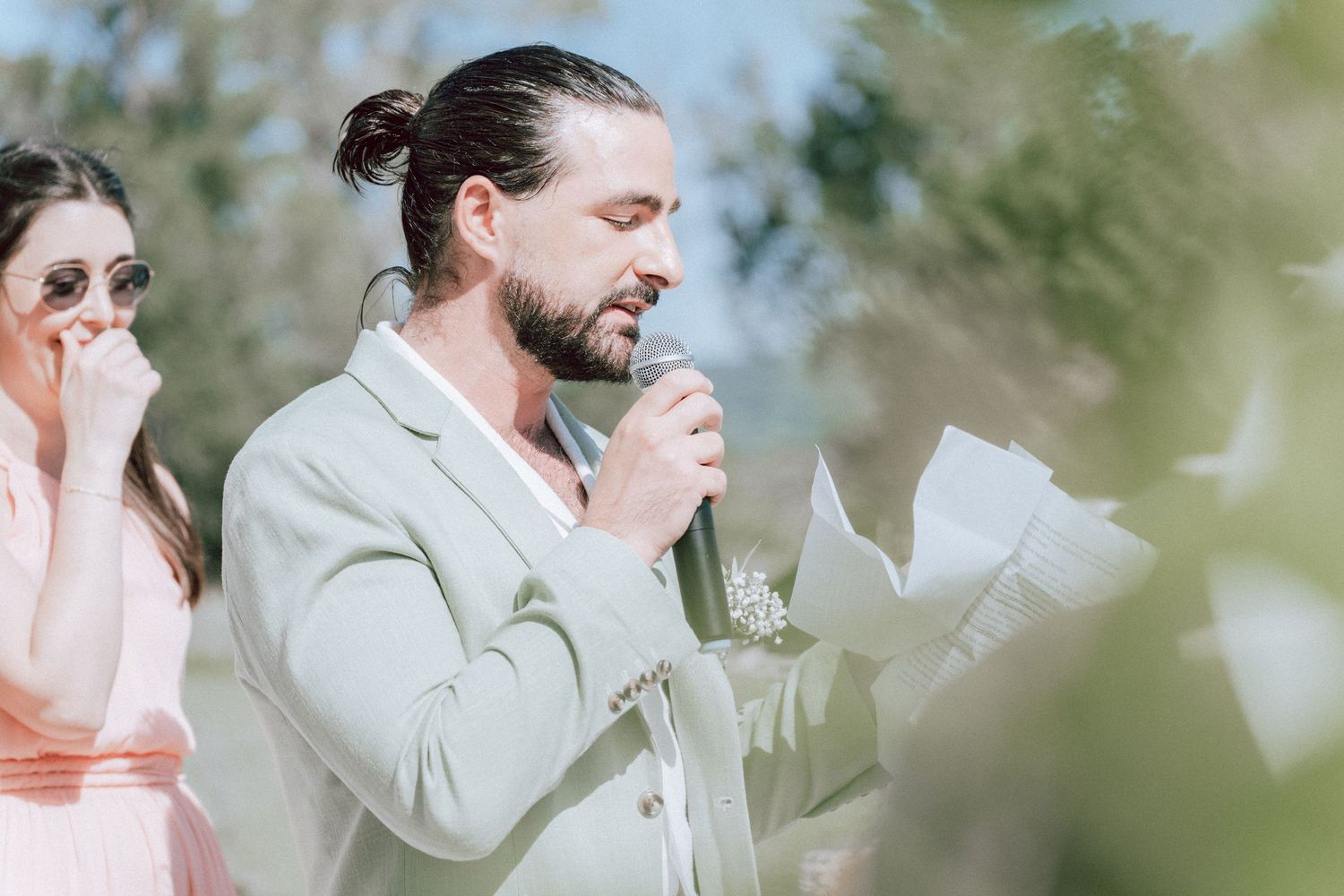 Man in sage green suit reading vows into microphone at outdoor wedding ceremony.