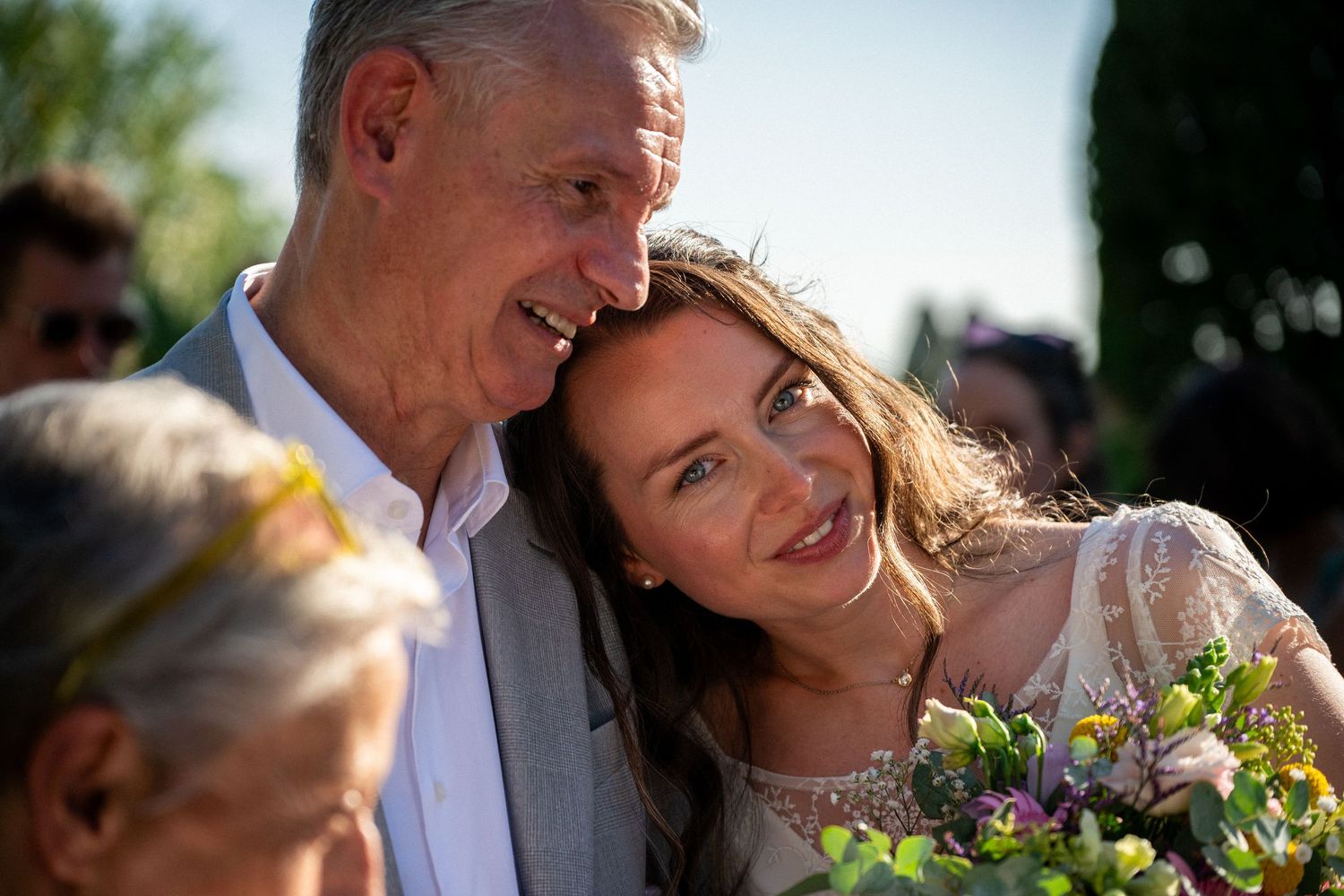 Bride smiling with older man at outdoor wedding ceremony, holding colorful flower bouquet in warm sunlight.