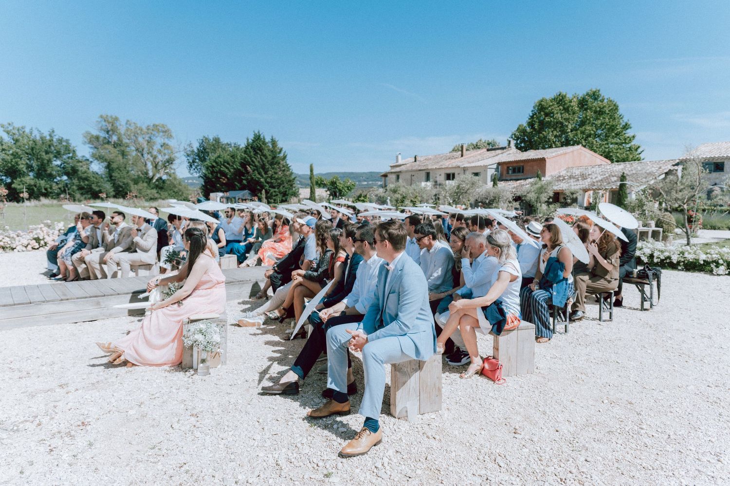 Wedding guests seated outdoors in sunny Provence countryside ceremony venue.