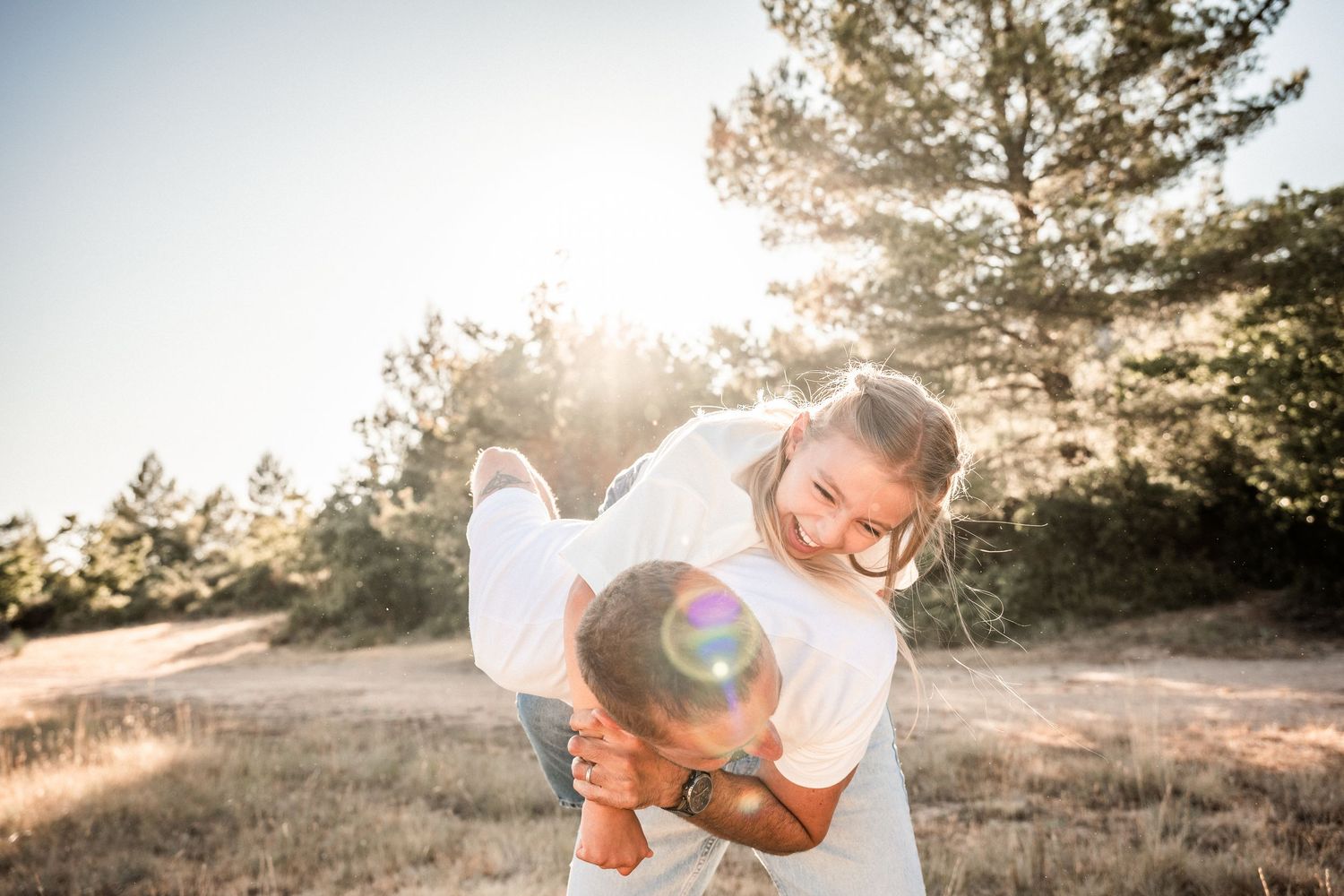 Couple laughing outdoors in golden sunlight, man lifting woman over his shoulder in a sunny meadow.