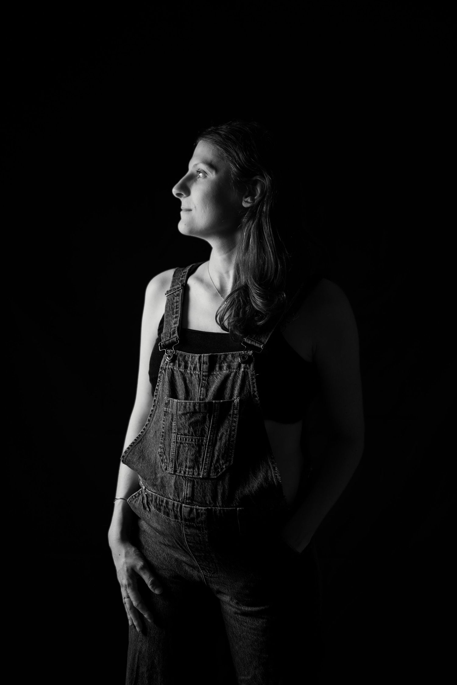 Pregnant woman in denim overalls looking upward in dramatic low-key black and white studio portrait.