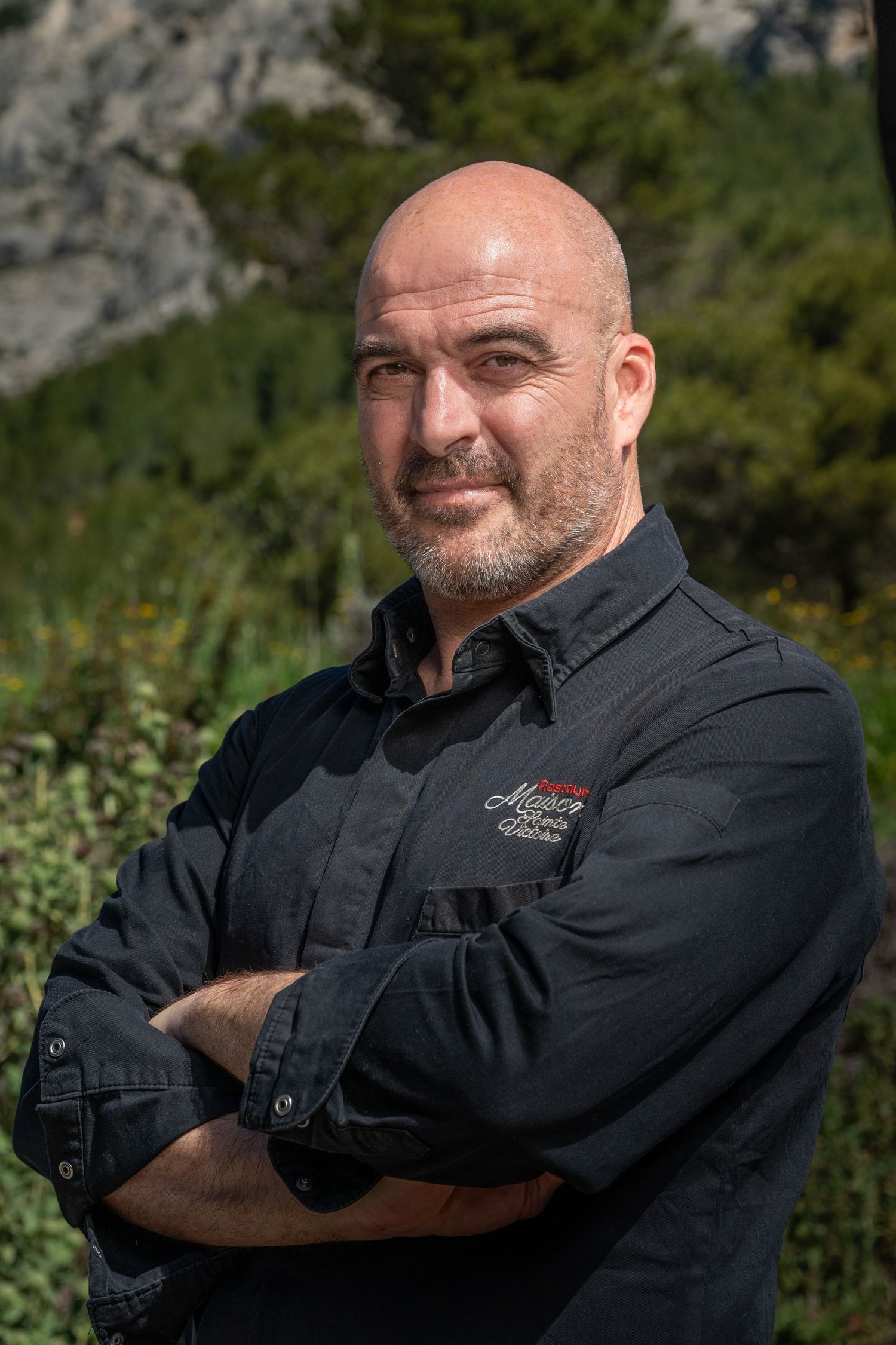 Confident bald man with beard in black shirt standing with arms crossed outdoors.