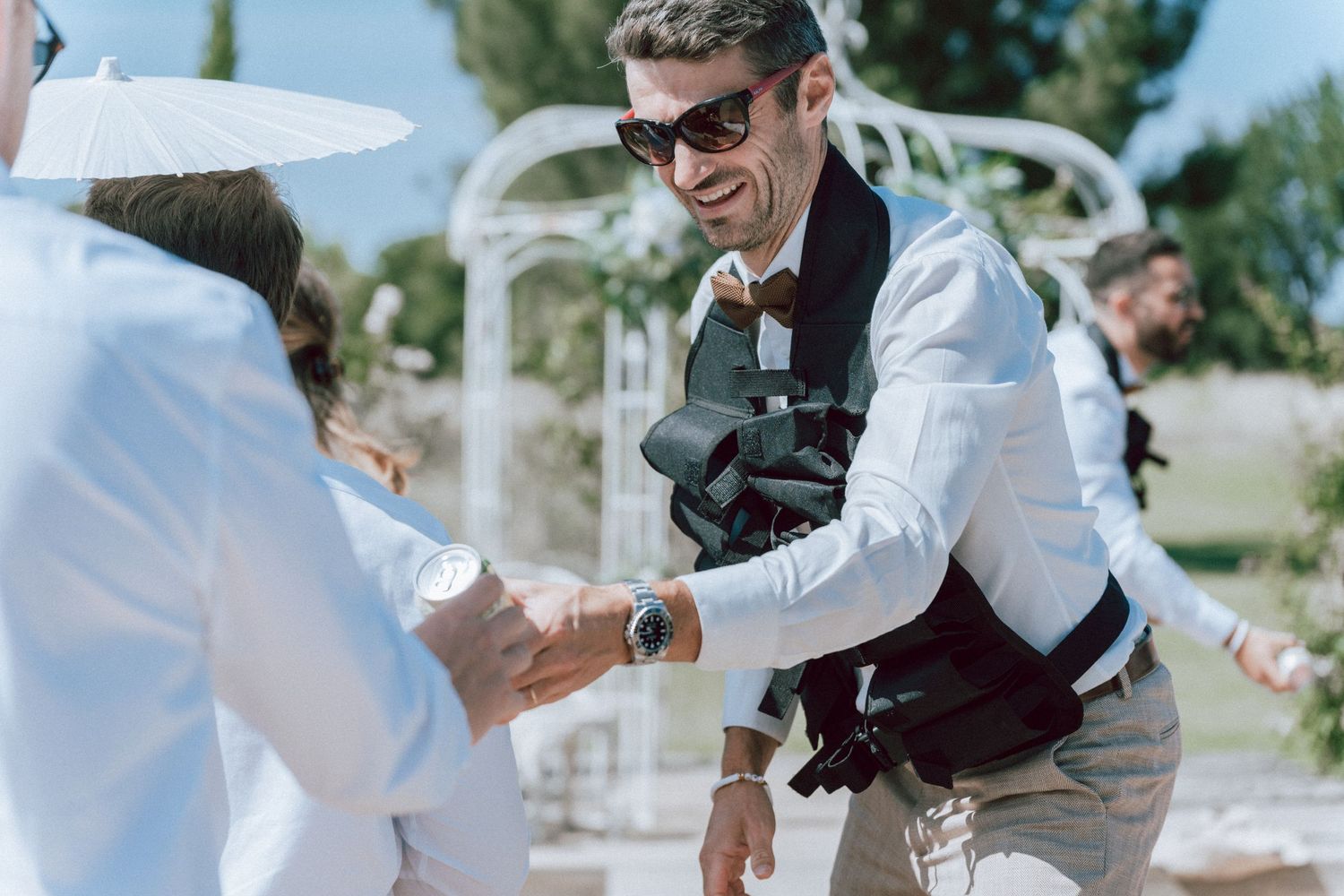 Smiling wedding photographer in sunglasses and camera vest shaking hands with guest at outdoor ceremony.