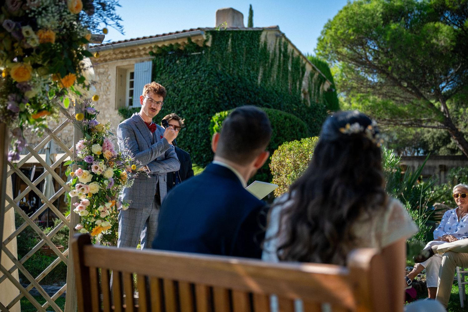 Officiant speaks at outdoor wedding ceremony with couple seated on wooden bench amid floral decorations.