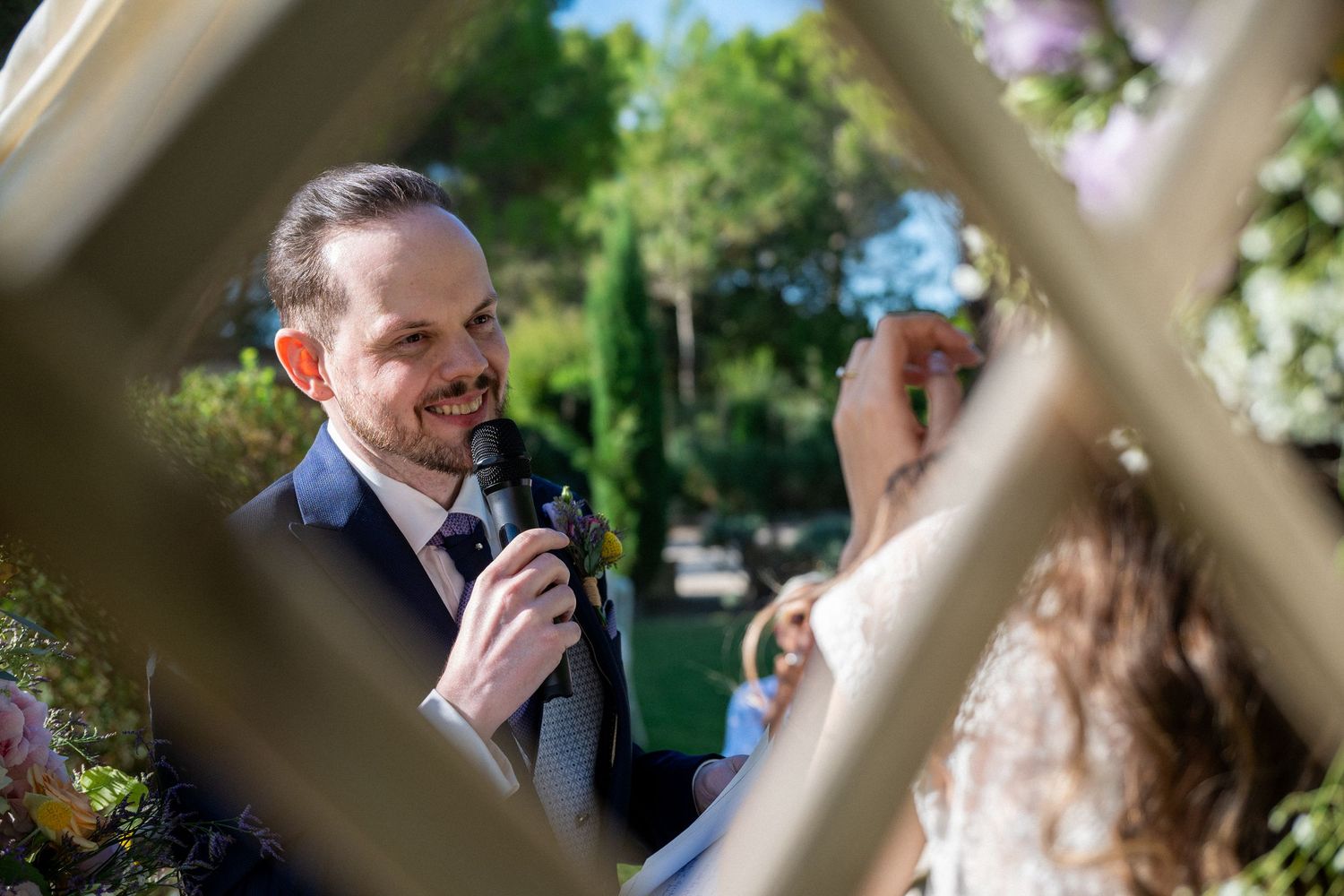 Groom smiling and speaking into microphone during outdoor wedding ceremony.