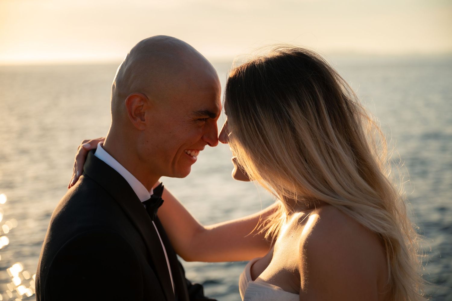 Bride and groom touching foreheads romantically with golden sunset ocean backdrop.