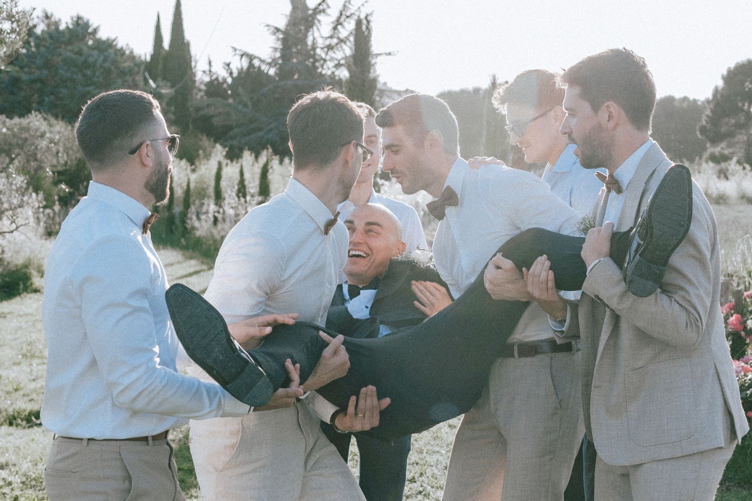 Groomsmen joyfully lifting groom in dark suit during outdoor wedding celebration in garden.