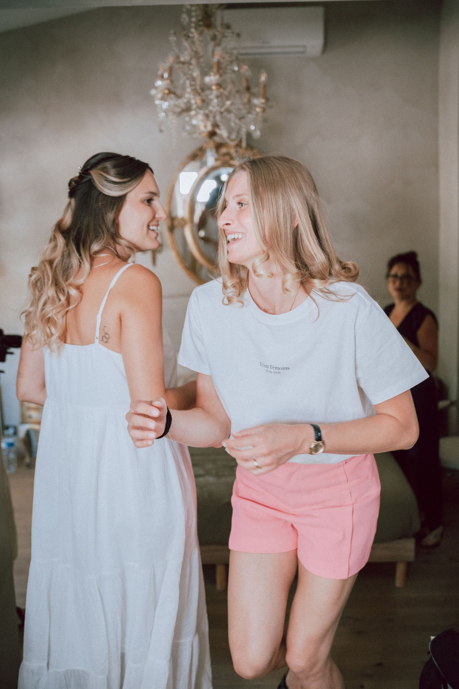 Two bridesmaids laugh and dance together in a bright room during wedding preparations.