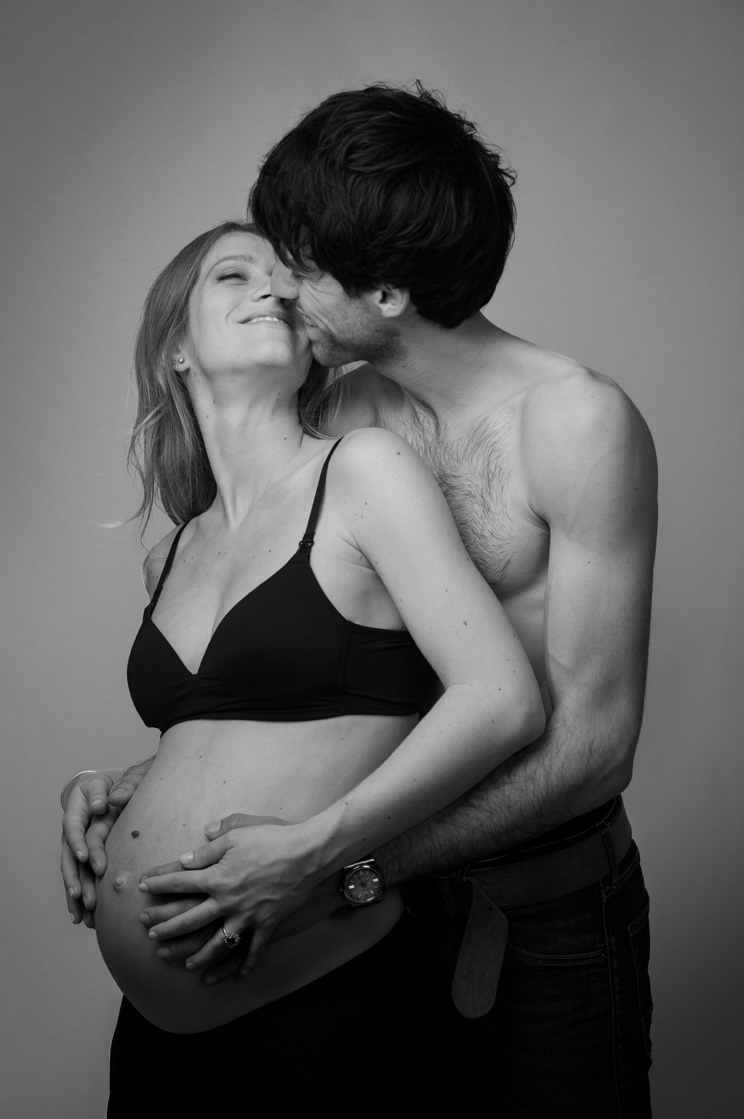 Black and white photo of shirtless man tenderly kissing pregnant partners cheek.