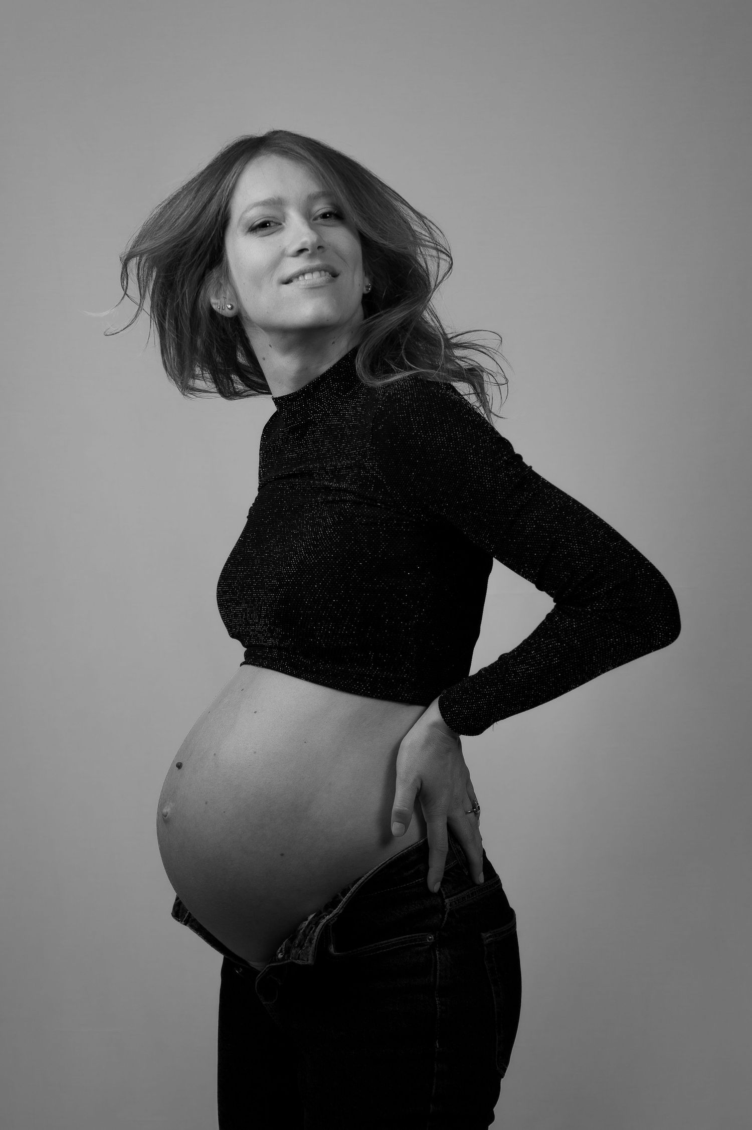 Smiling pregnant woman in black crop top and jeans showing baby bump in black and white studio portrait.