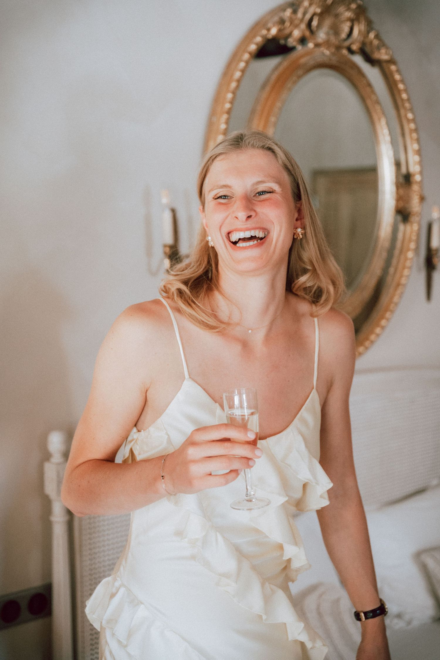 Joyful bride laughing in white ruffled dress holding champagne glass before ornate gold mirror.