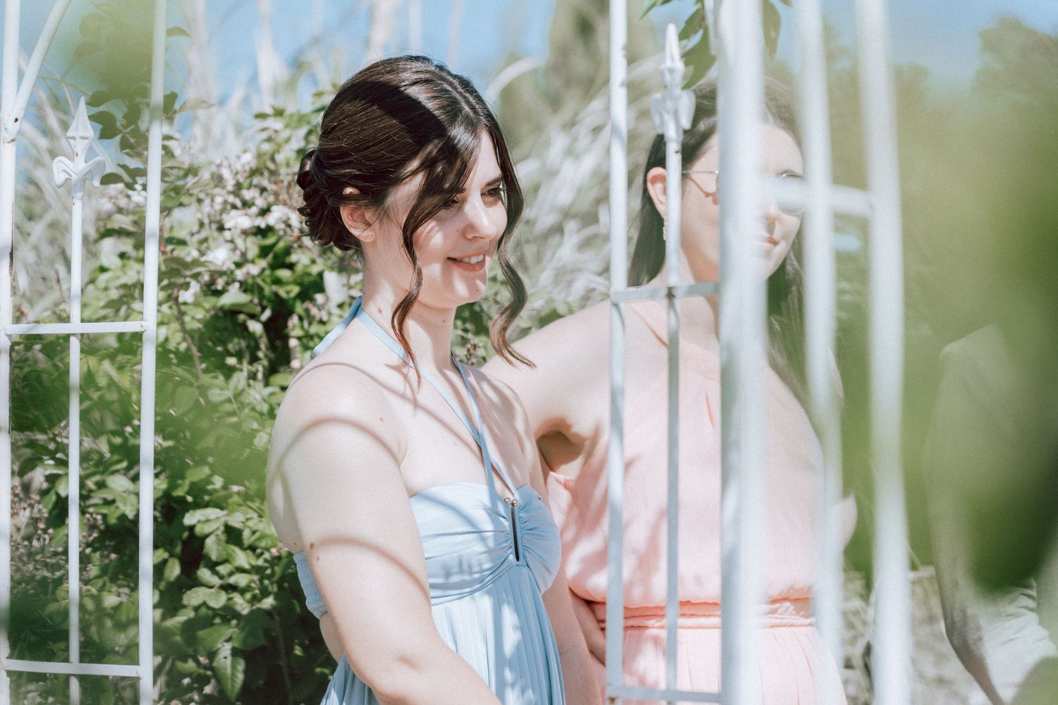Two bridesmaids in pastel dresses smile near a white garden gate surrounded by lush greenery.