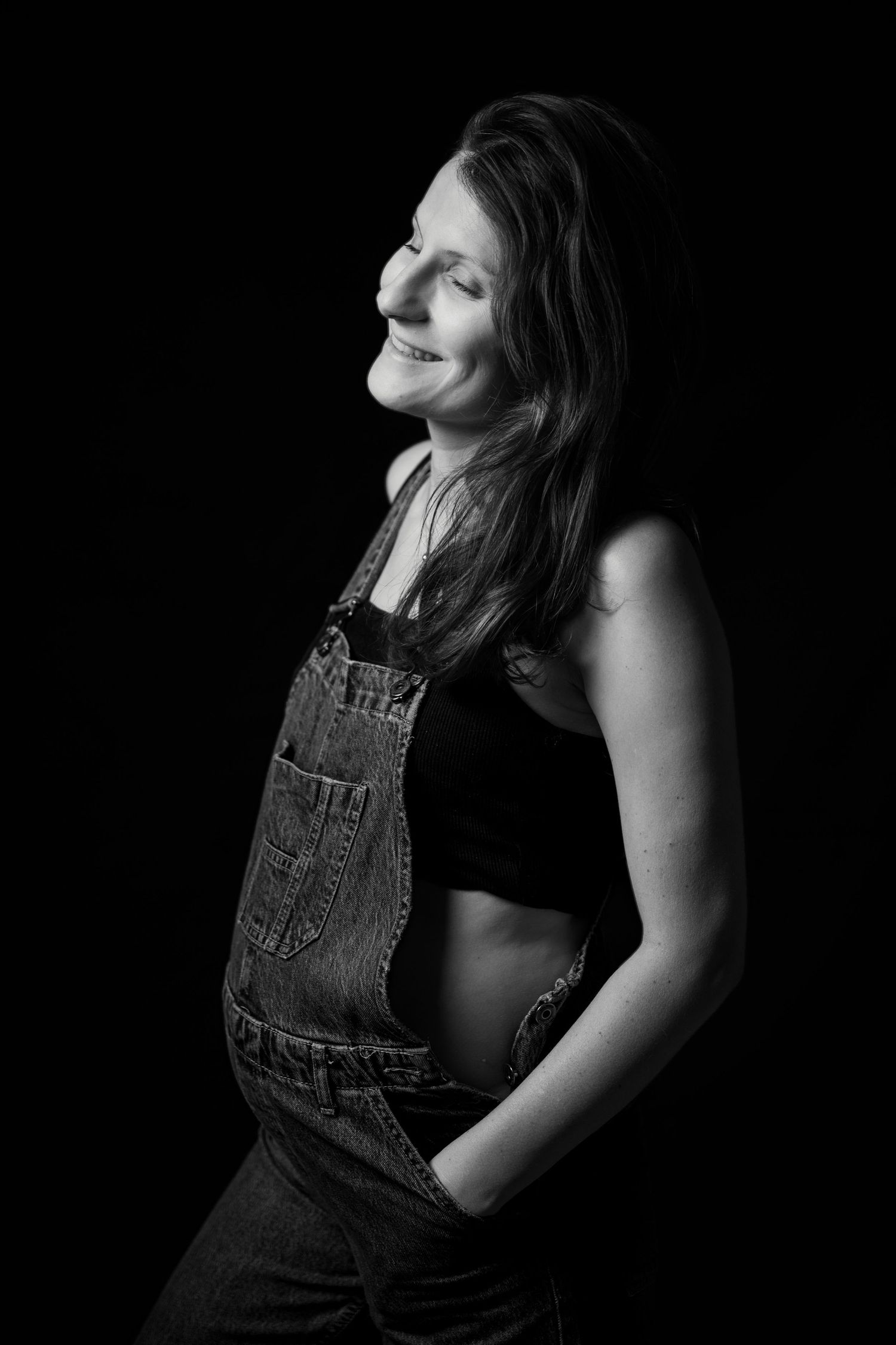 Black and white studio portrait of a smiling pregnant woman in denim overalls and crop top against dark background.