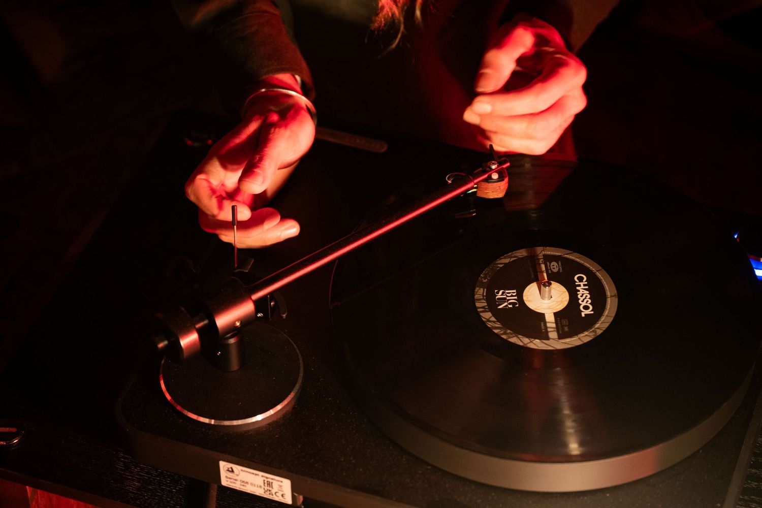 DJ hands adjusting a vinyl record needle on a turntable in dark club lighting.