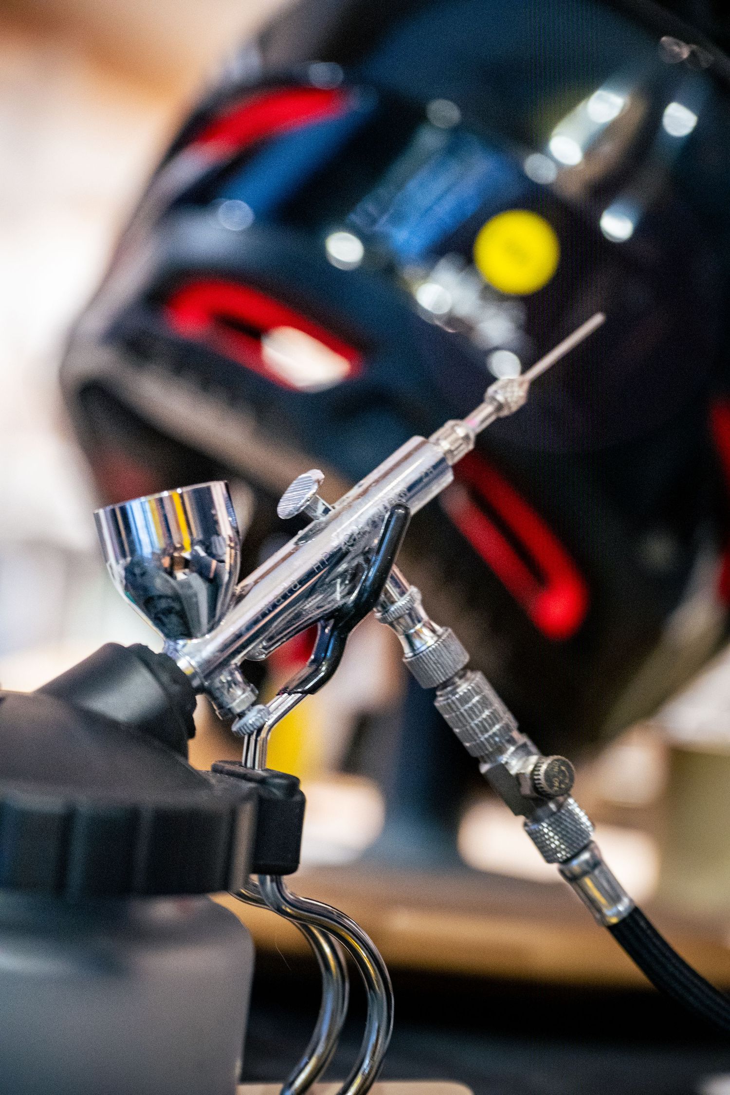 Close-up of a chrome airbrush gun held by a gloved hand, with a blurred black car in the background.