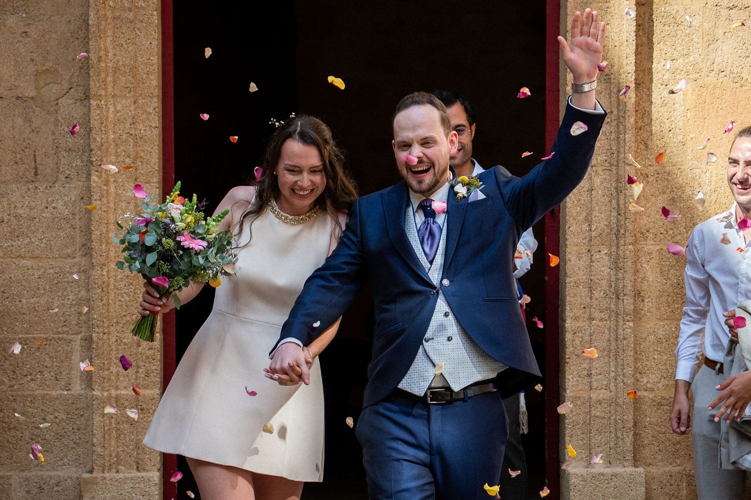 Newlywed couple exits church in confetti shower, bride holding bouquet, groom waving in navy suit.