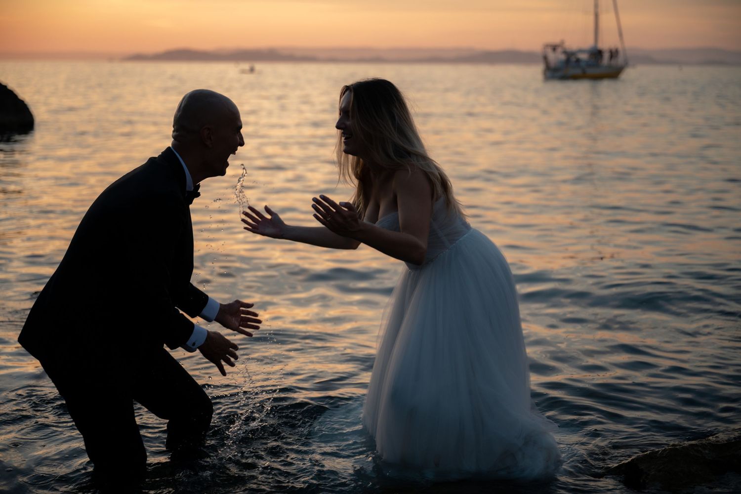 Playful newlyweds splash water at each other on rocky beach during stunning sunset.