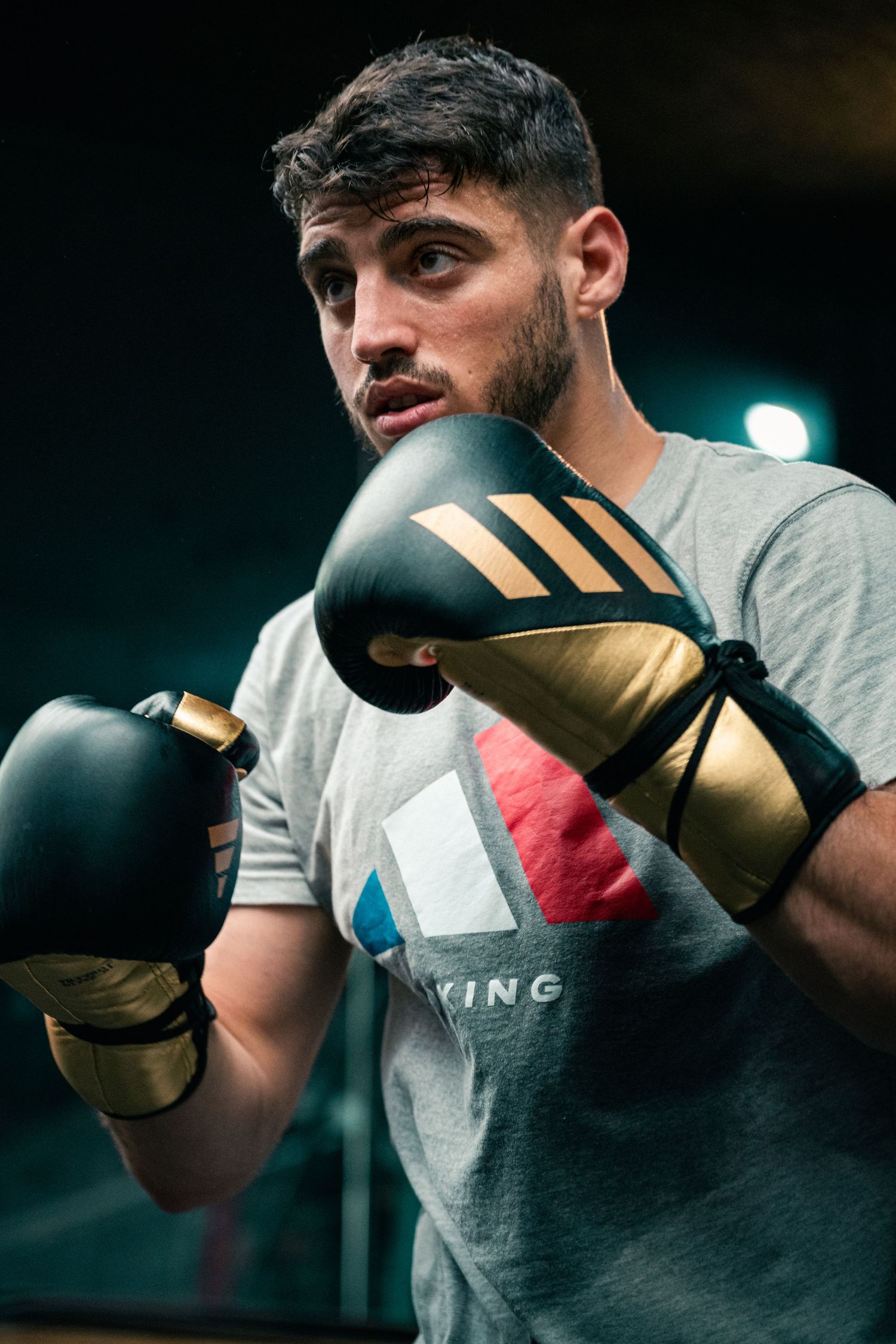 Male boxer in Adidas gloves training in fighting stance in a dark gym setting.