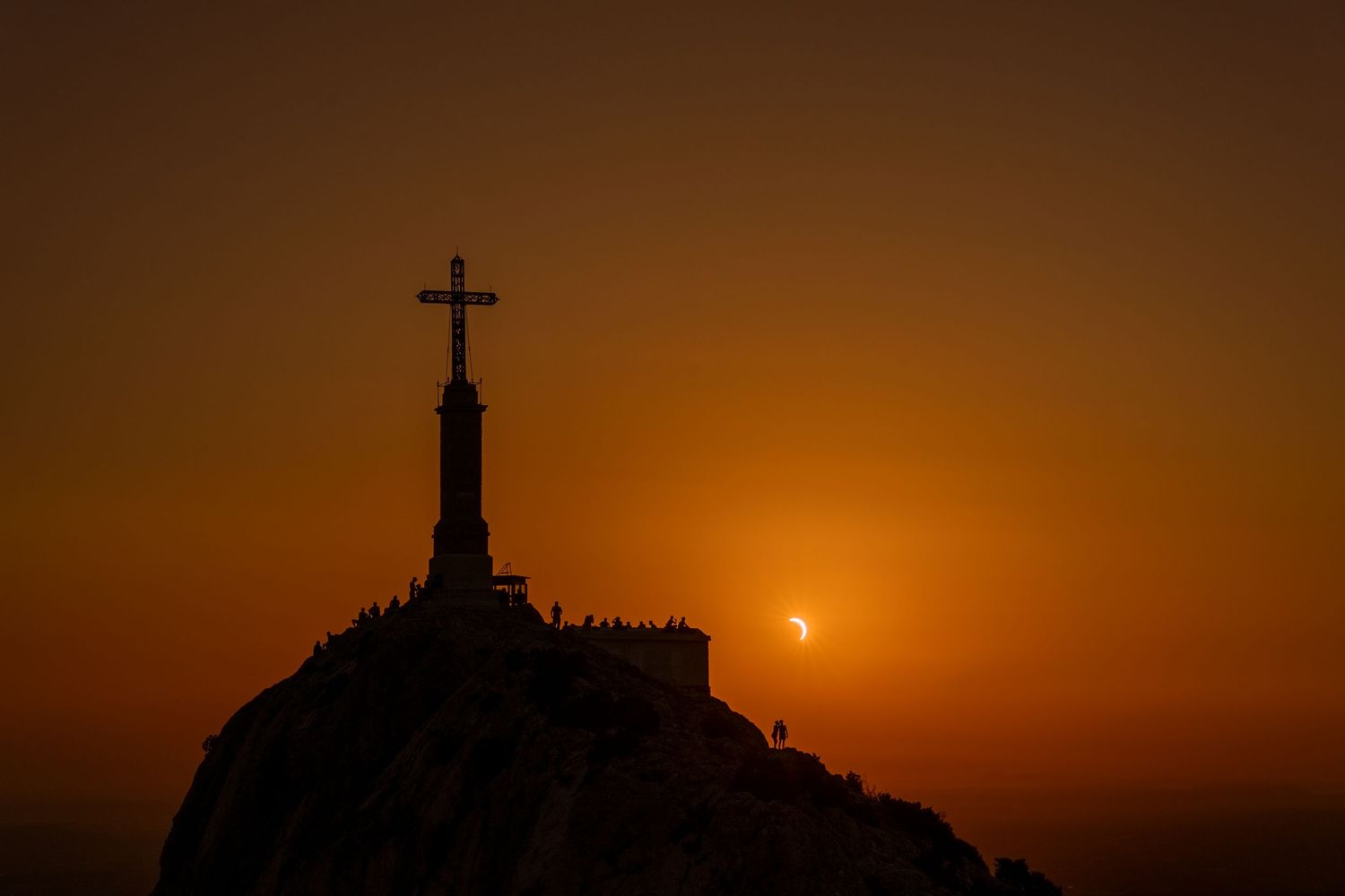 Silhouette of a cross atop a rocky hill against a vibrant orange sunset sky.
