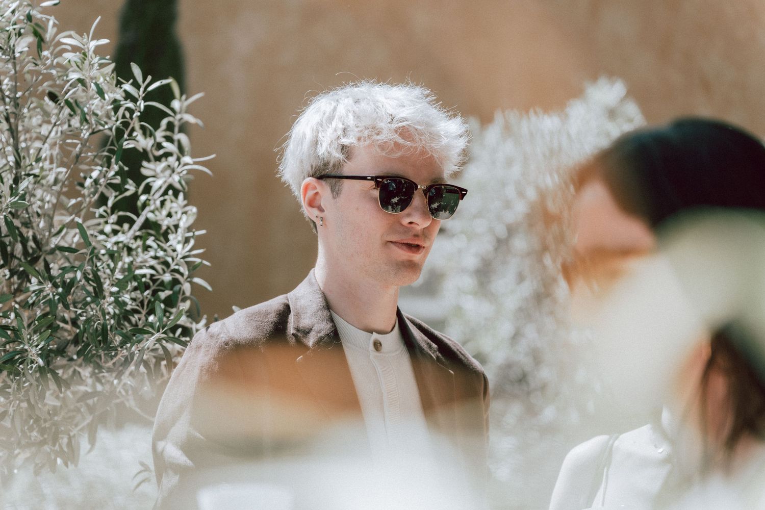 Young man with white hair and sunglasses poses outdoors among lush green plants in natural light.