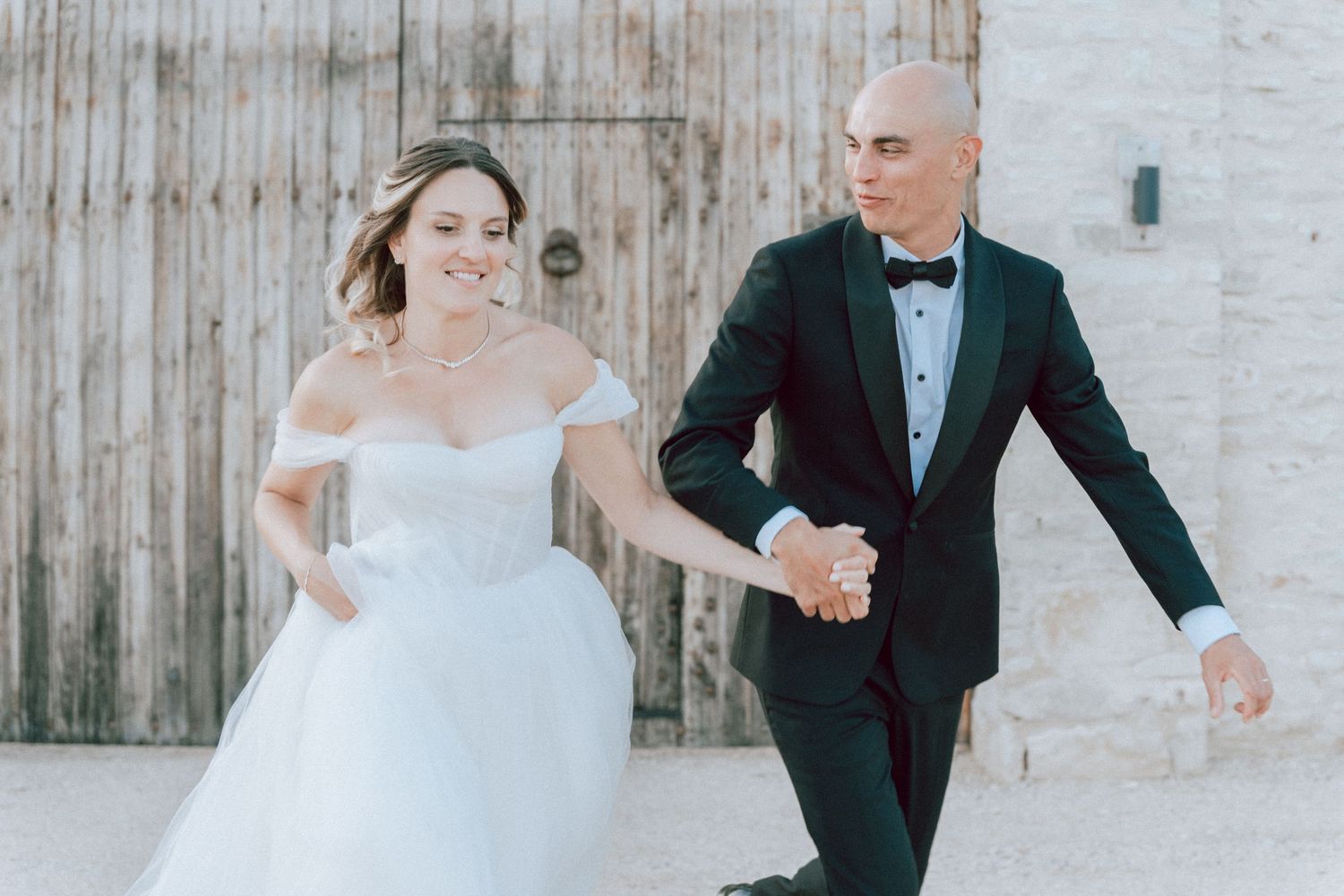 Bride and groom hold hands and walk together in front of a rustic wooden barn door.