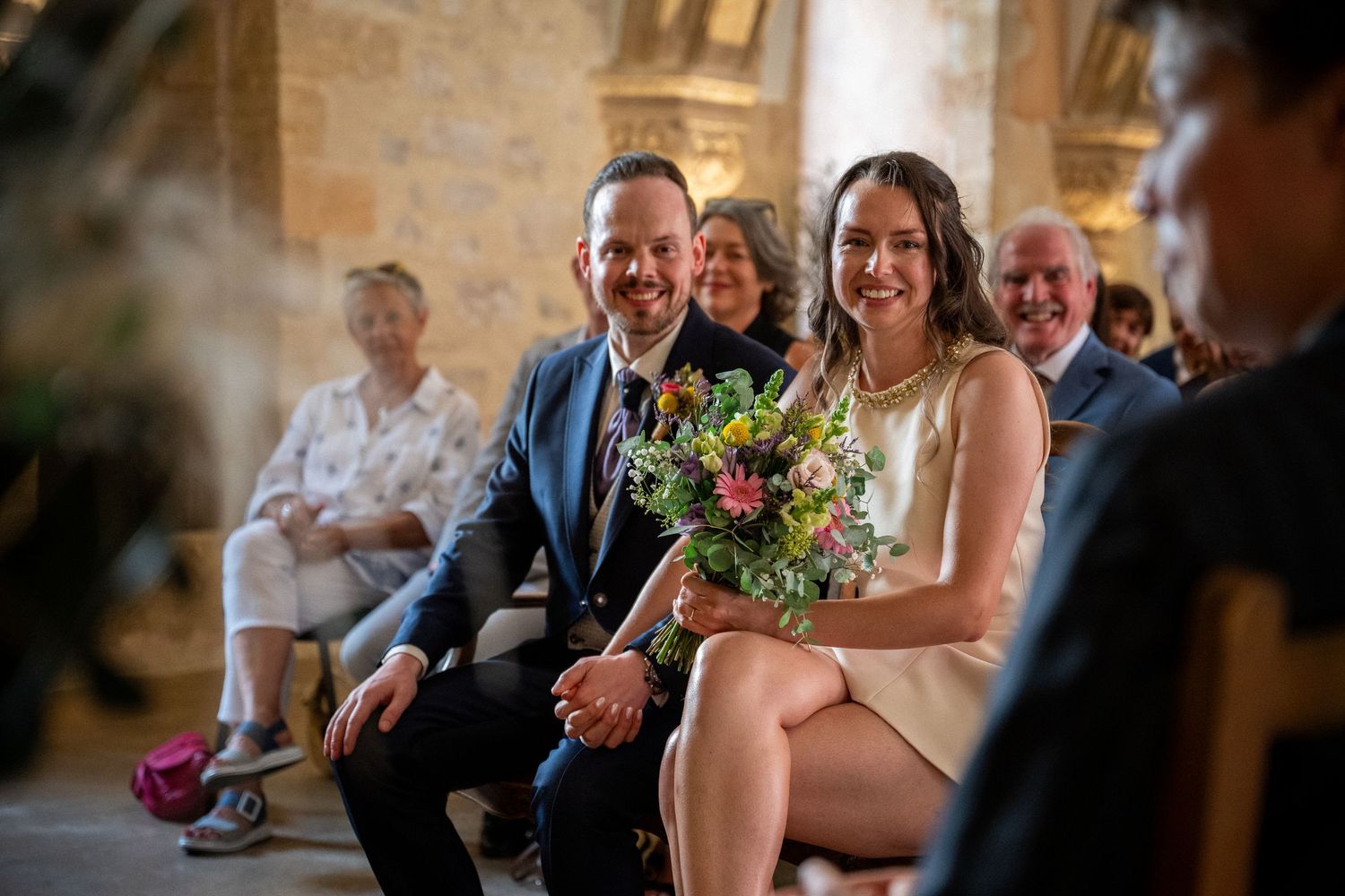 Happy couple sits together holding a bouquet during a wedding ceremony in a rustic stone venue with guests smiling behind them.