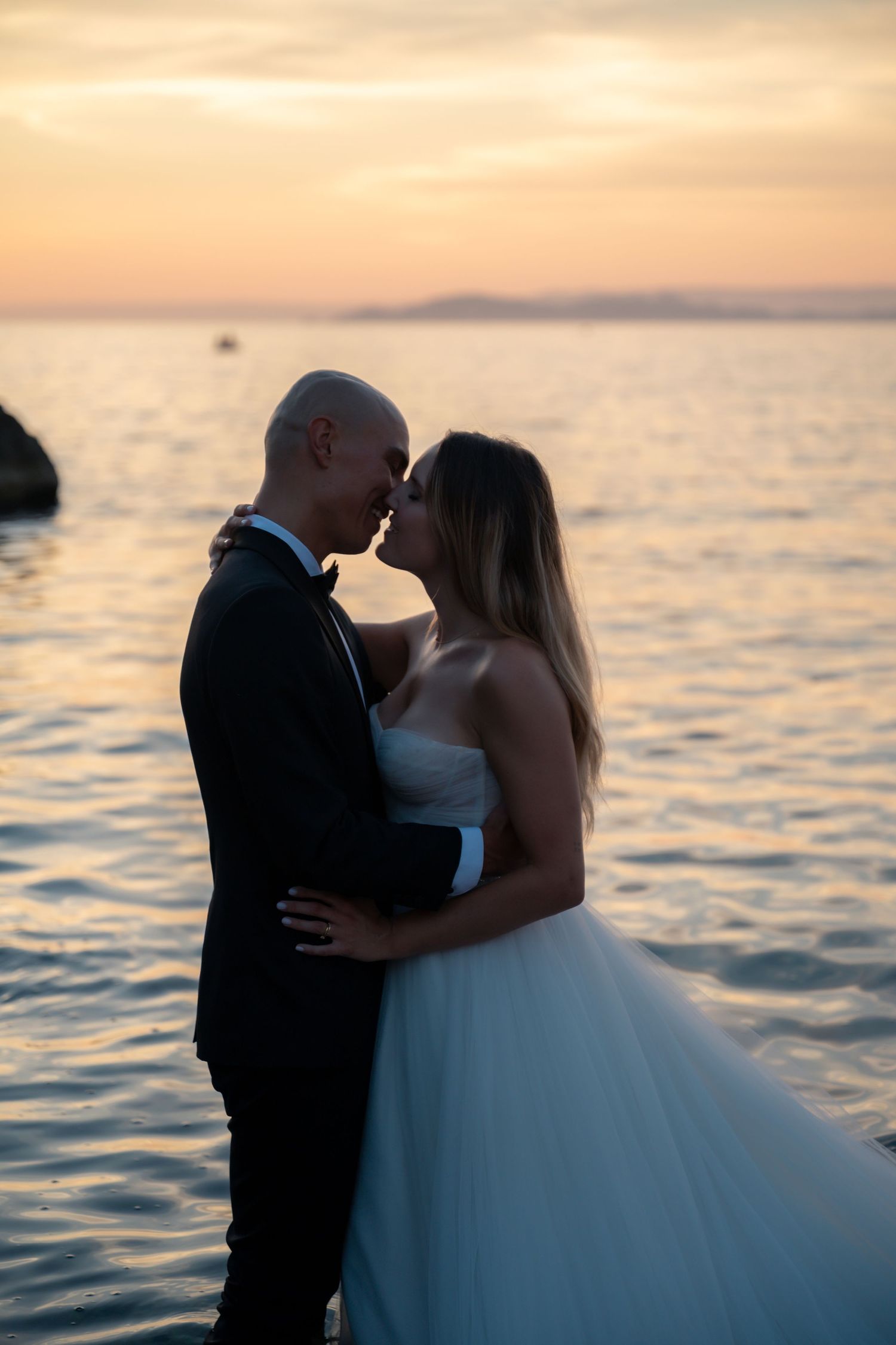 Romantic bride and groom nearly kissing silhouetted against vibrant orange sunset over calm sea.