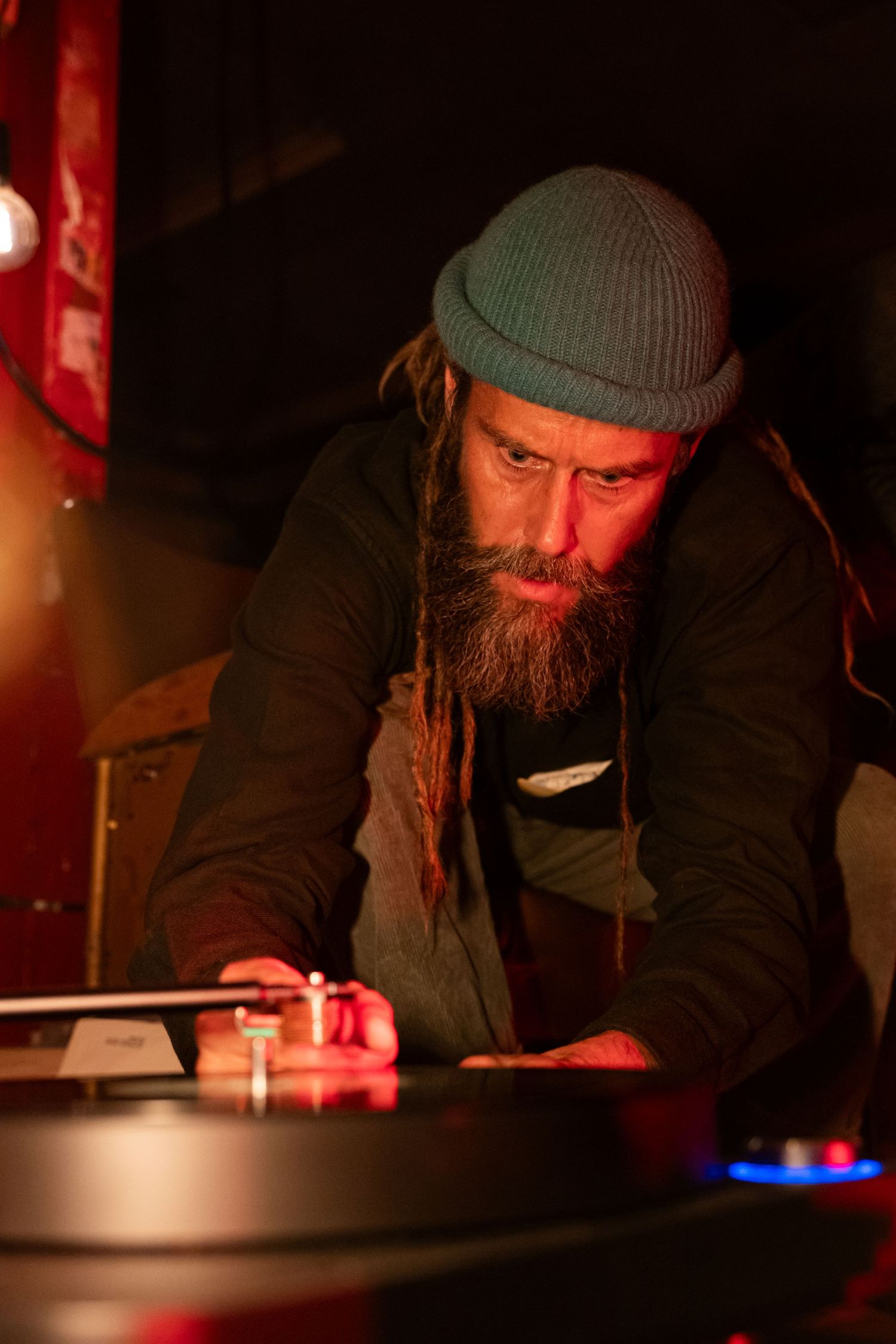 Bearded man with dreadlocks and blue beanie hat focused on DJ turntables in dark red-lit venue.