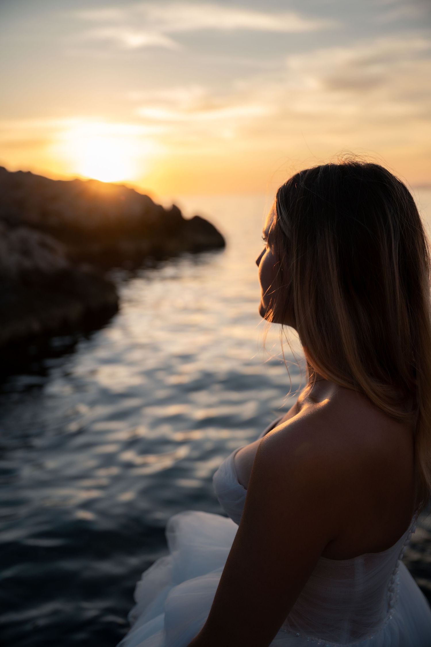 Bride in white gown gazing at golden sunset over calm rocky coastline.