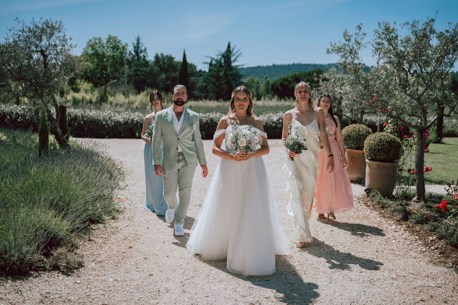 Bride in white gown walks with wedding party down a gravel path surrounded by lavender and greenery.