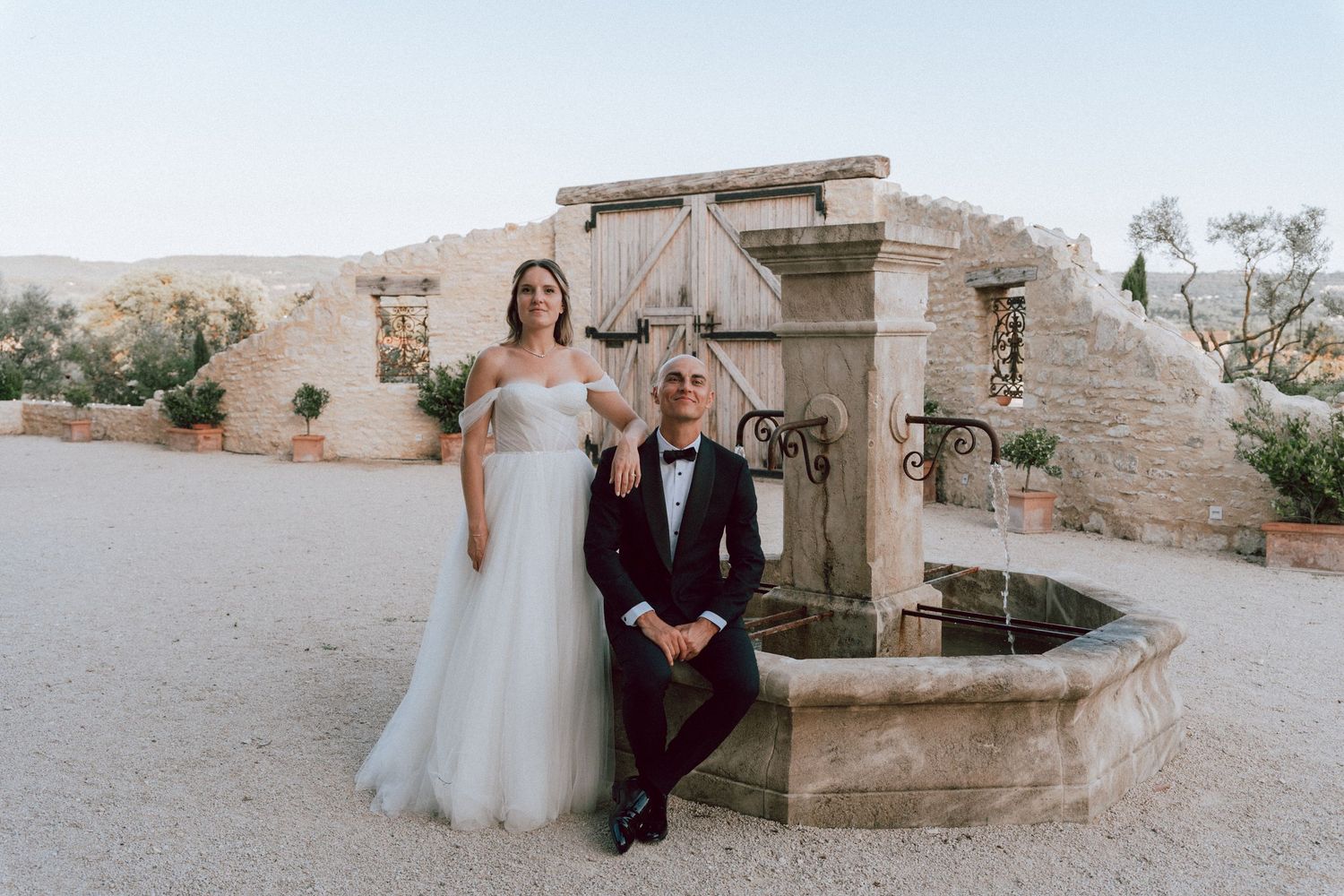 Bride in white off-shoulder gown and groom in black tuxedo posing by stone fountain at rustic venue.
