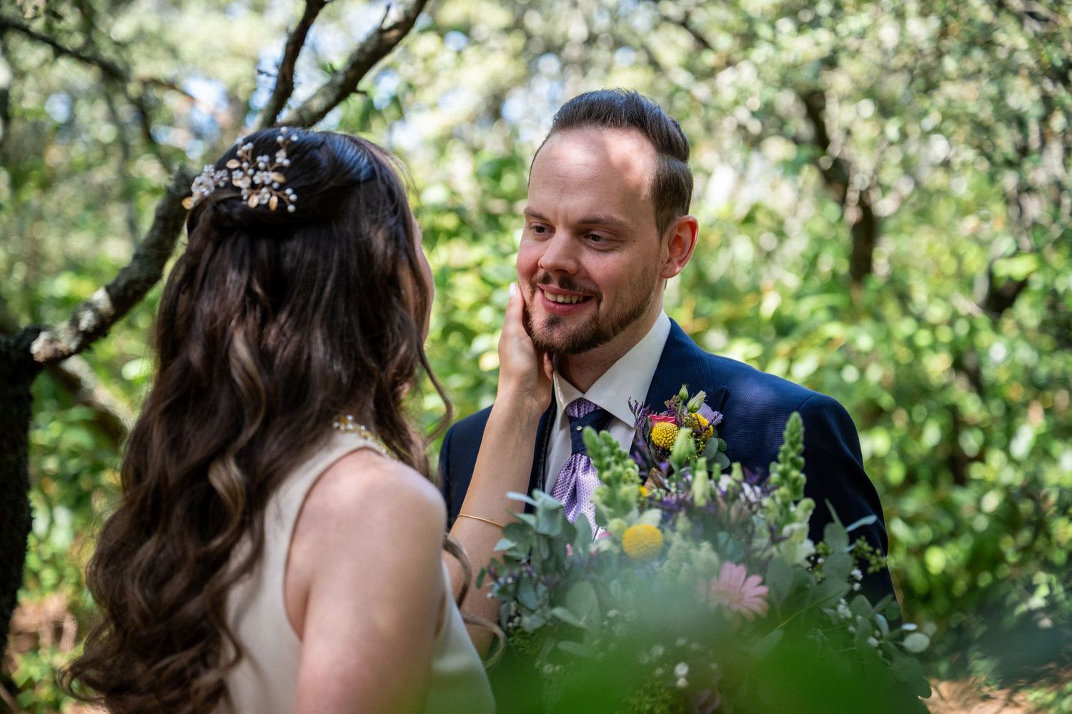 Bride tenderly touches groom's face in lush garden setting, both smiling during romantic outdoor wedding ceremony.