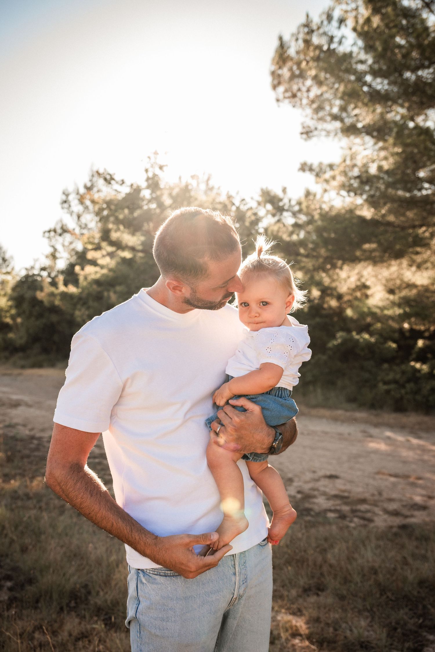 Father kissing baby outdoors in golden sunlight, holding infant in white outfit among green trees.
