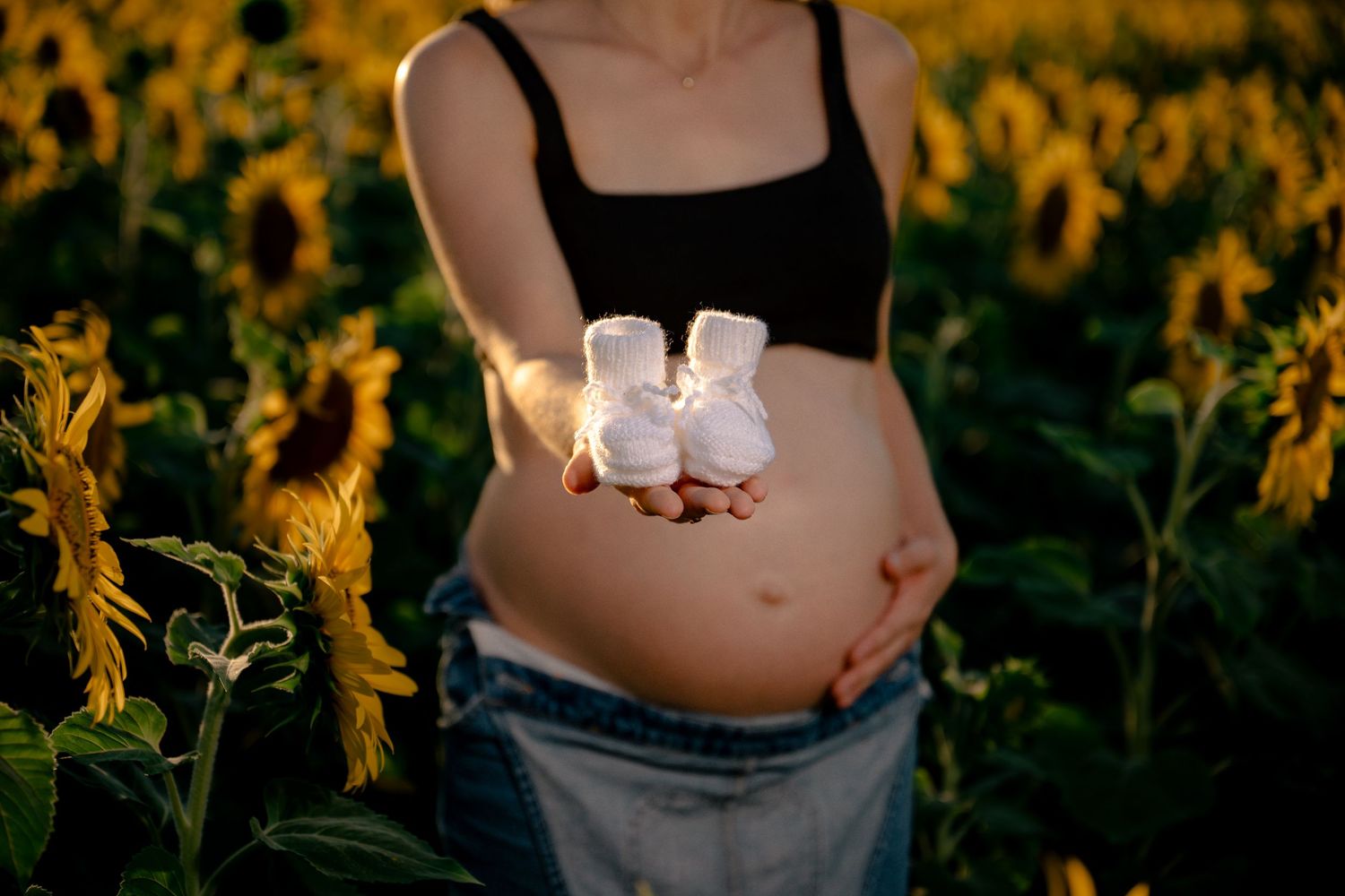 Pregnant woman holding tiny white baby booties in front of her bump in a sunflower field at sunset.