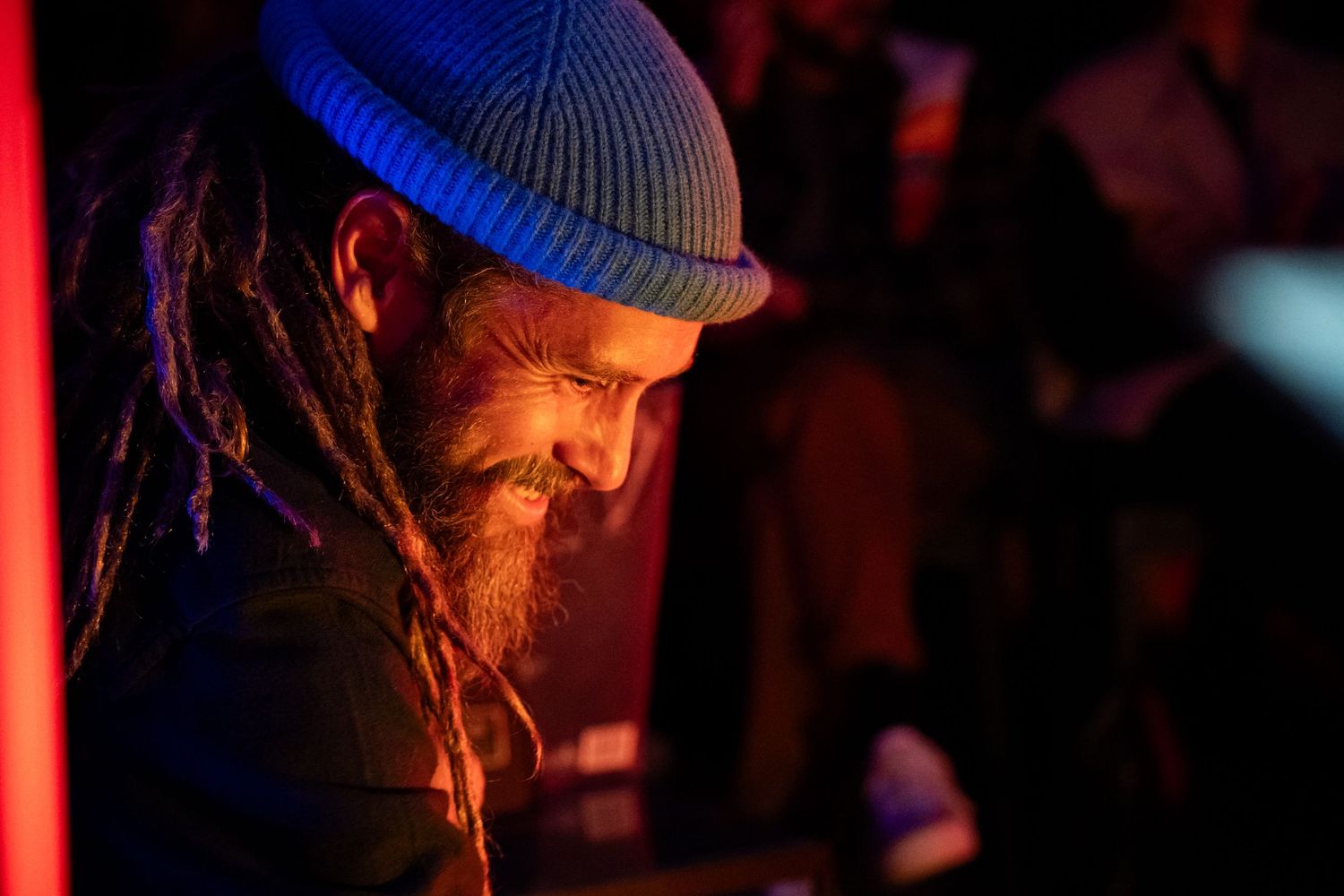 Bearded man with dreadlocks and blue beanie hat smiling under warm red and orange stage lighting.