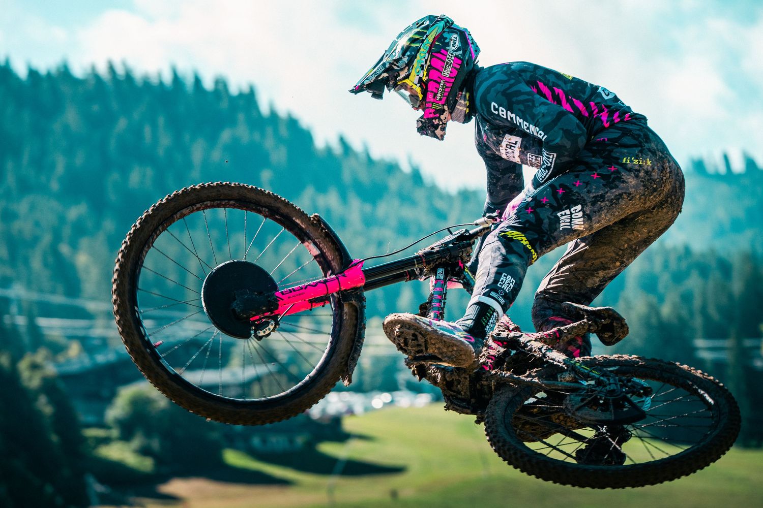 Muddy mountain biker performs aerial trick on pink bike wearing colorful helmet at downhill MTB race event.