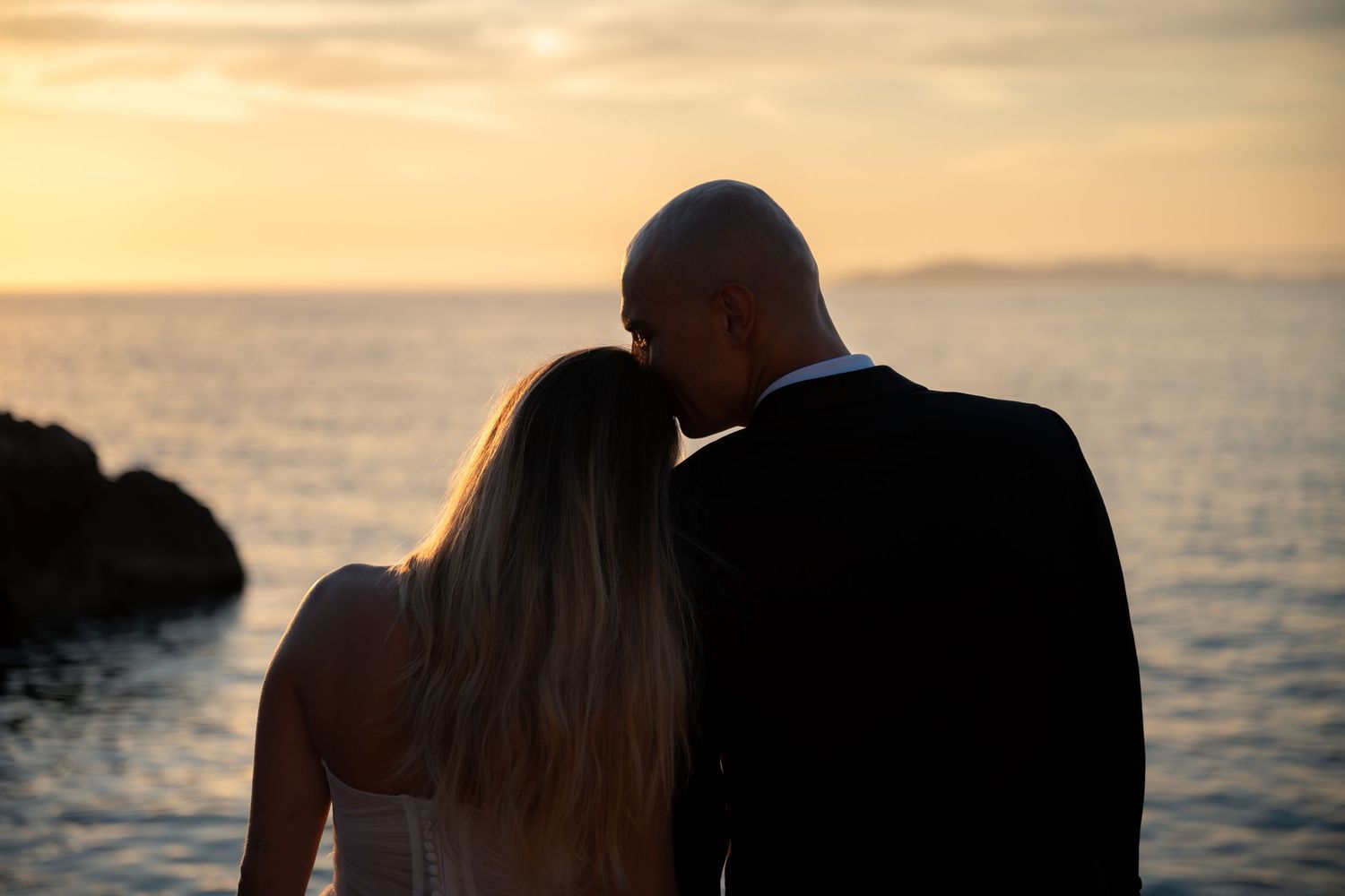 Couple sharing a romantic moment by the ocean at sunset, bride in white dress and groom in dark suit.
