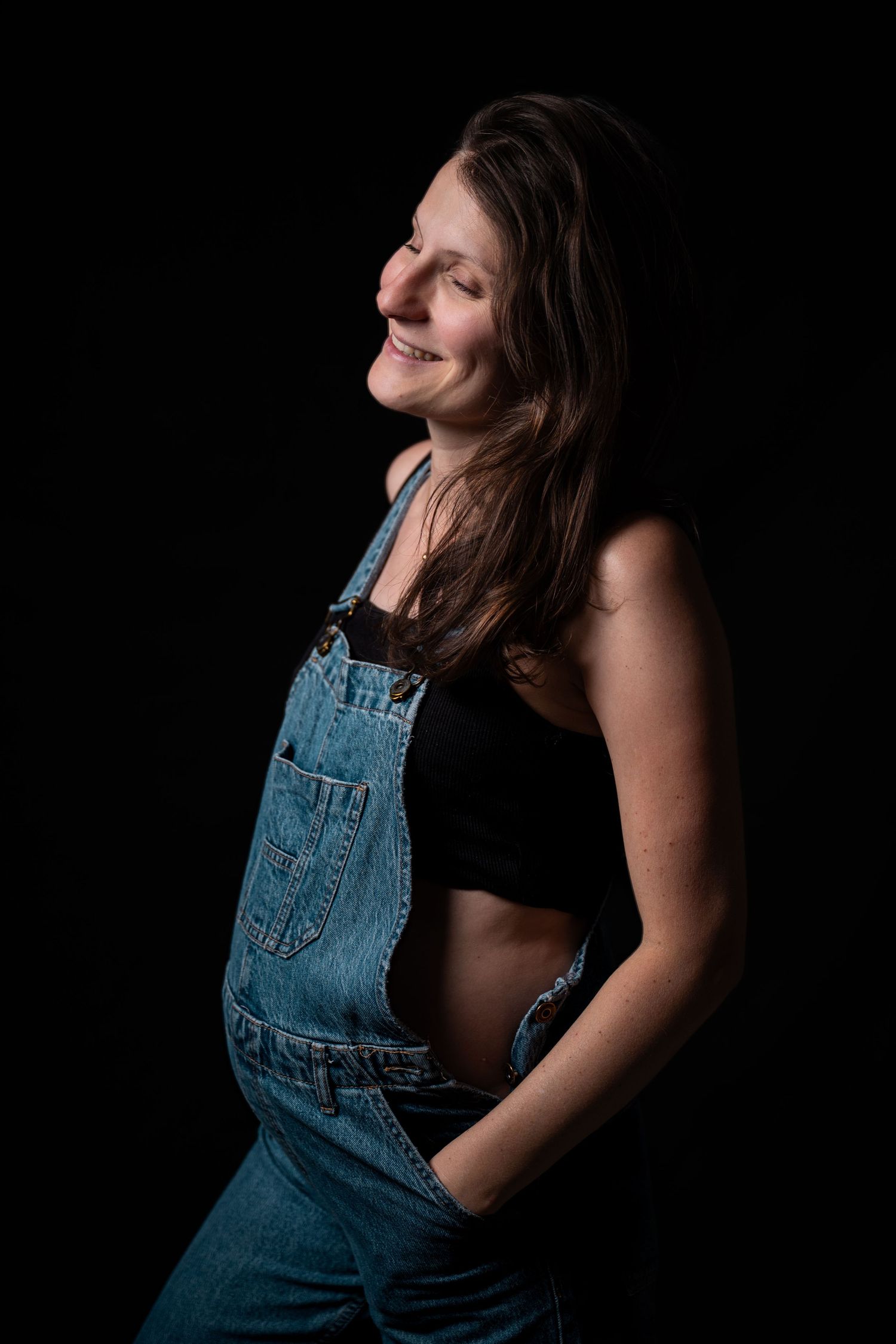 Smiling young woman in denim overalls and black crop top posing against dark background.